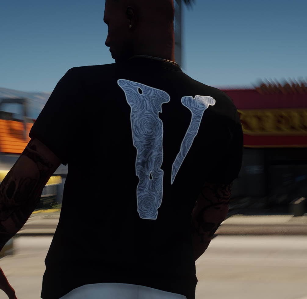 Vlone 桌布 壁紙 — 一名身穿黑色T恤、背後有一個大型風格化「V」字的男子，站在陽光普照的城市街道上，手臂上的紋身清晰可見。