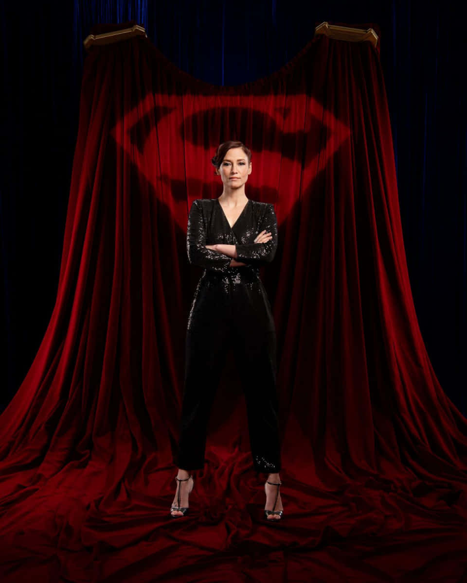 Chyler Leigh 桌布 壁紙 — 一名身穿黑色亮片連身褲的女性自信地交叉雙臂，站在紅色天鵝絨窗簾前，窗簾後顯現出超人標誌的投影。