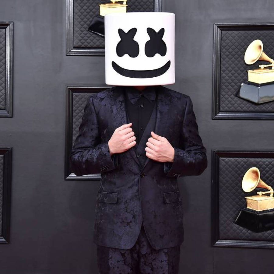 marshmello iPhone 桌布 壁紙 — 一名身穿黑色圖案西裝的人調整外套，戴著白色棉花糖面具（黑色眼睛和微笑），背景是裝飾著裱框葛萊美獎的深色牆壁。