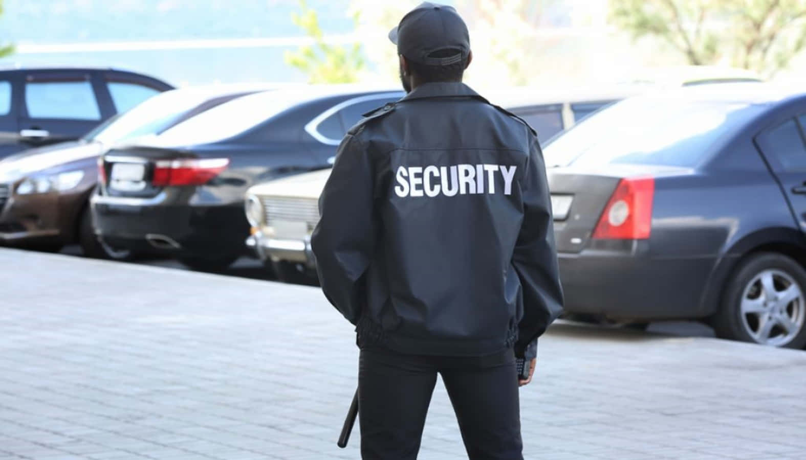 security 桌布 壁紙 — 一名身穿黑色外套、背後印有「SECURITY」字樣的保安員，在鋪砌的停車場中警覺地站立，周圍是汽車。
