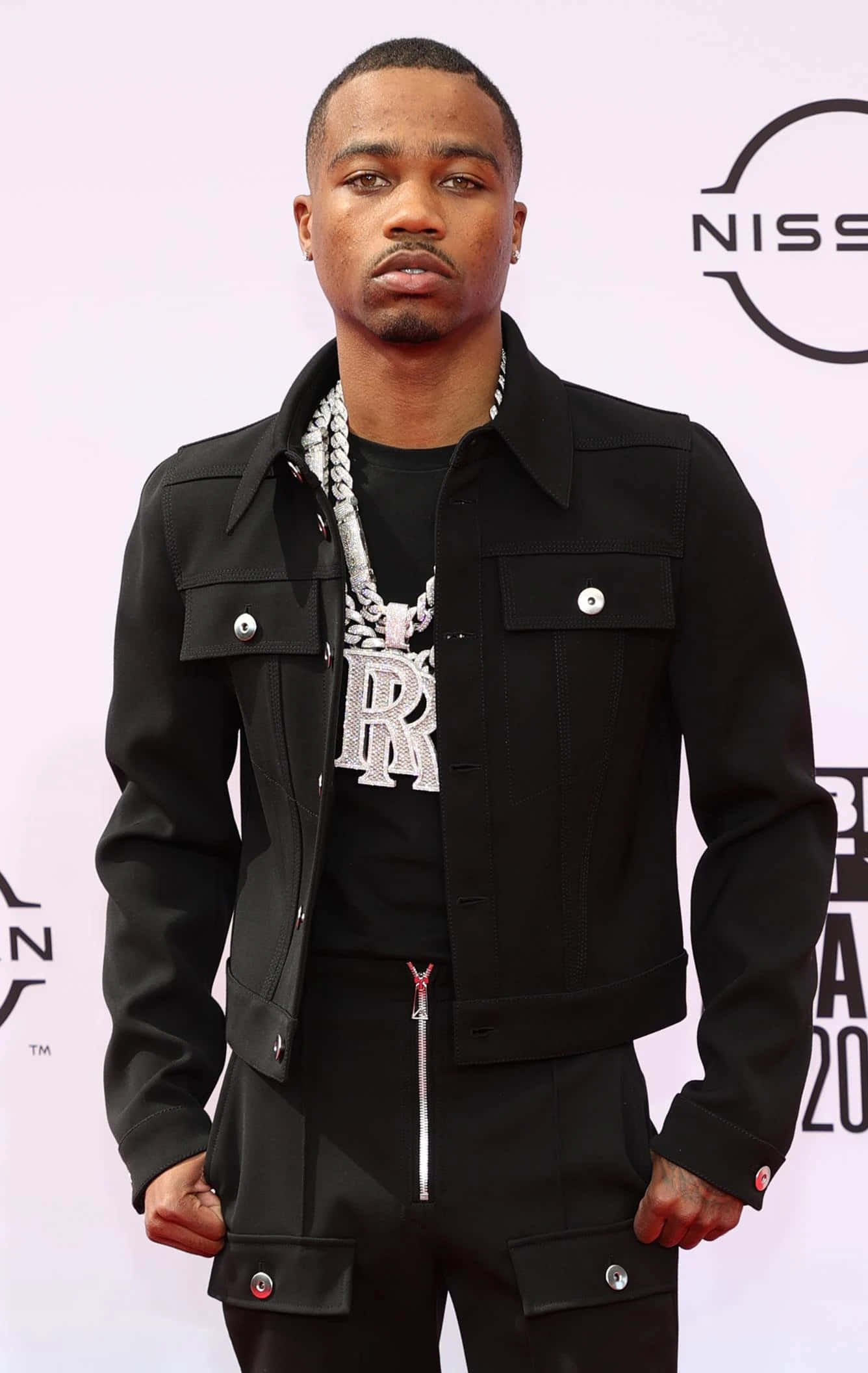 Roddy Ricch 桌布 壁紙 — 一名身穿黑色外套和褲子的男子自信地站在紅地毯上，戴著一條帶有「R」吊墜的大銀鏈。