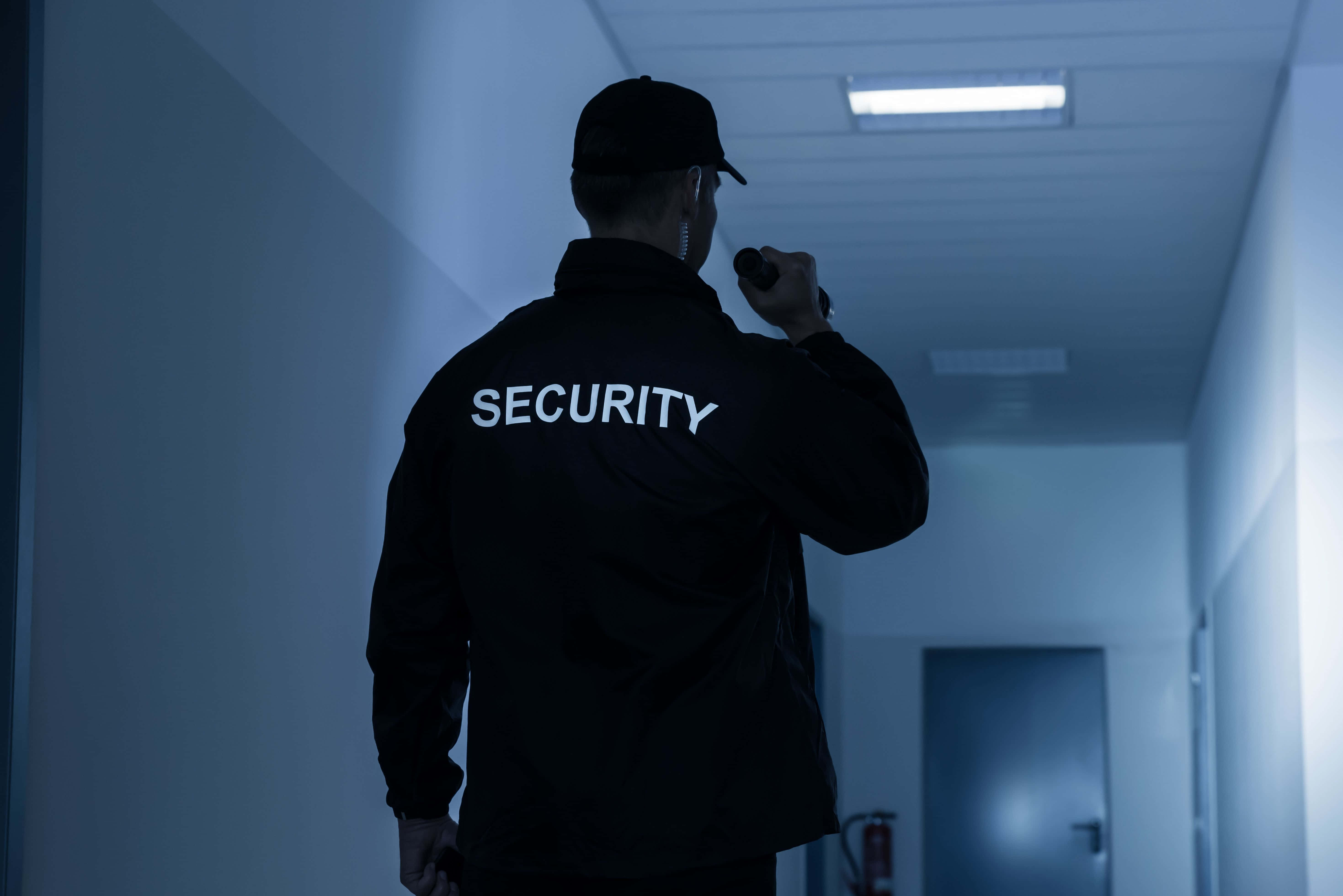 security 桌布 壁紙 — 一名身穿黑色外套的保安員手持電筒，沿著燈光昏暗的走廊行走。