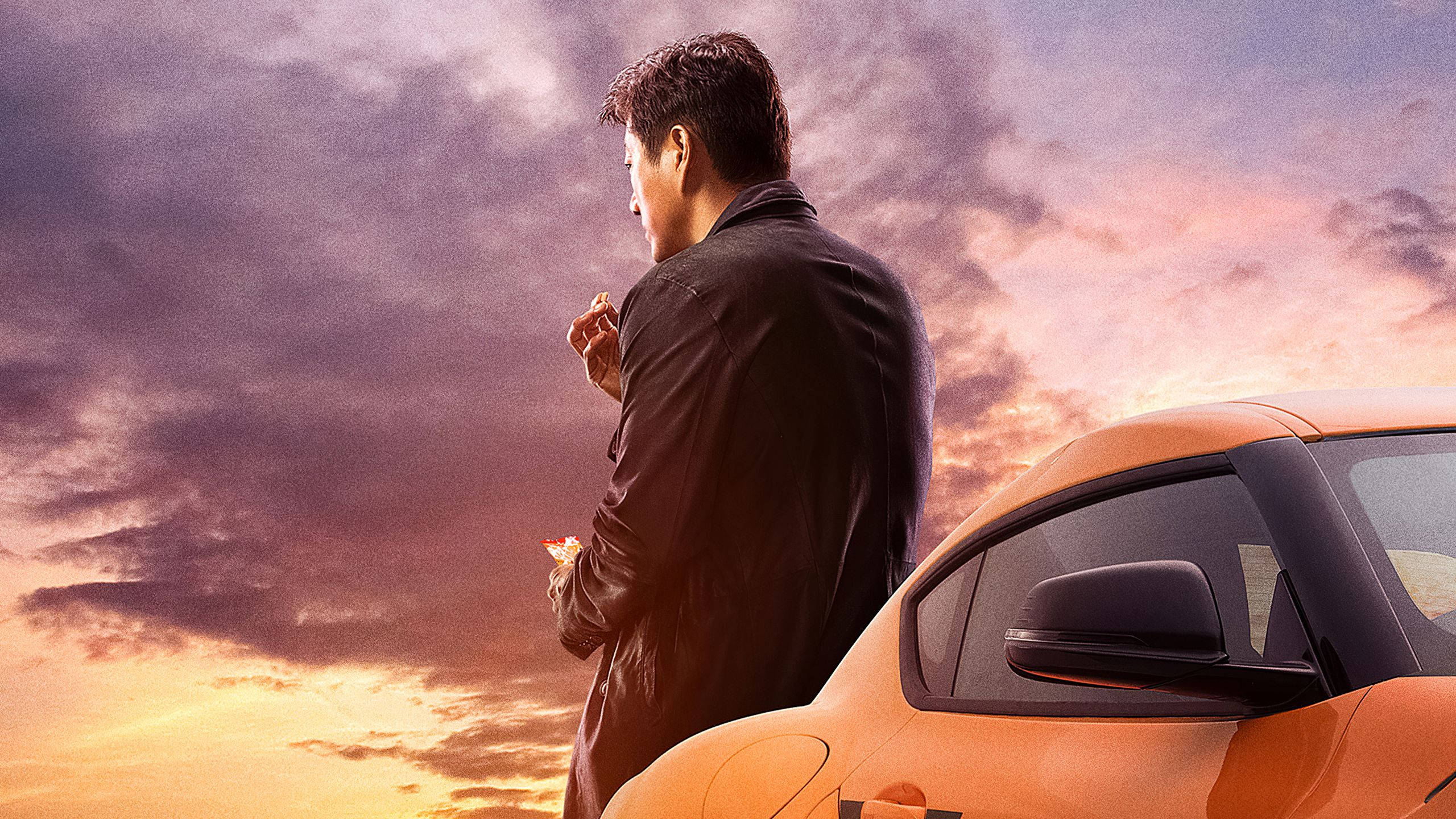Sports Car 桌布 壁紙 — 一名身穿黑色外套的男子站在橙色跑車旁，背景是紫色和金色雲彩的壯麗日落天空。