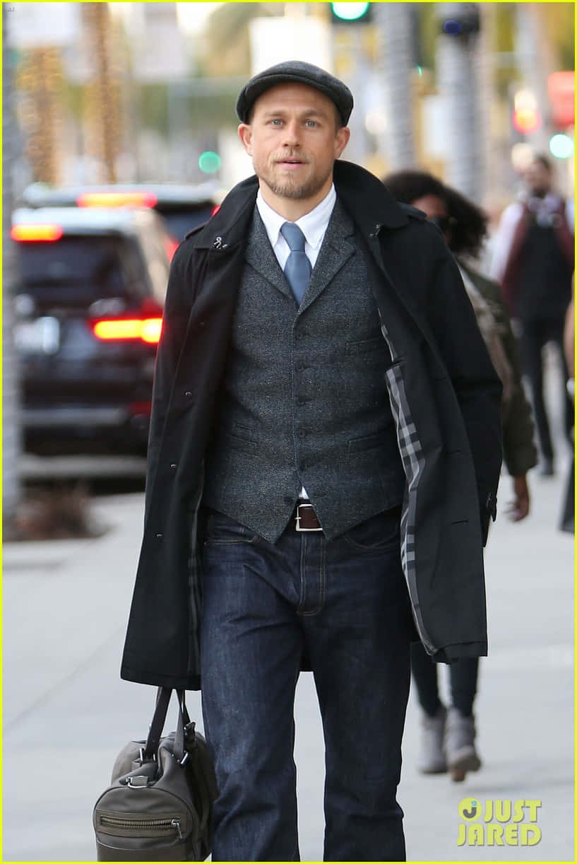 Dapper Charlie Hunnam 壁紙 — 一名身穿黑色外套背心和帽子的男子自信地走在城市人行道上，背景中汽車和行人模糊不清。