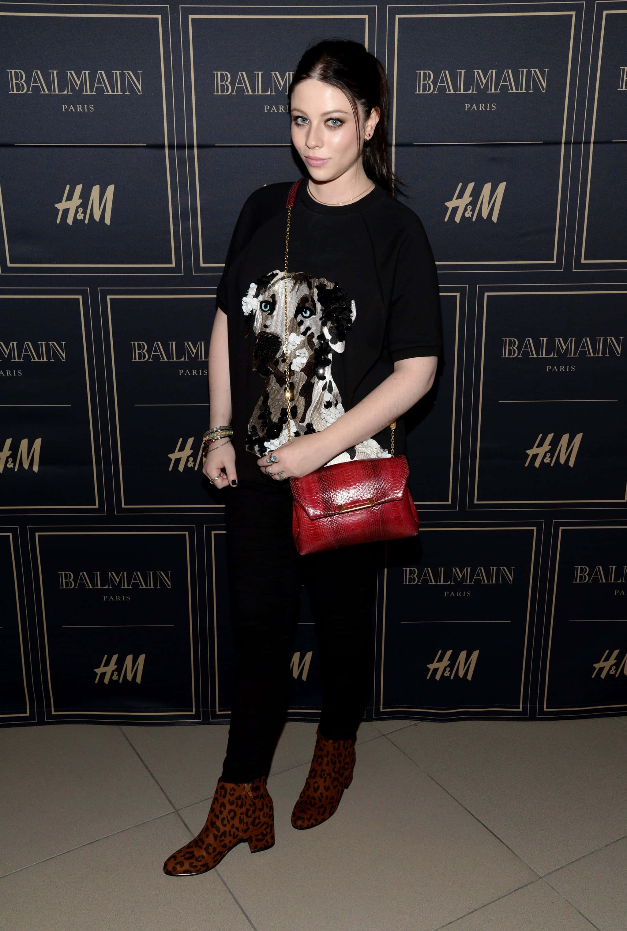 michelle trachtenberg 桌布 壁紙 — 一名身穿黑色服裝、豹紋靴和紅色手袋的女性在活動中站在品牌背景前。