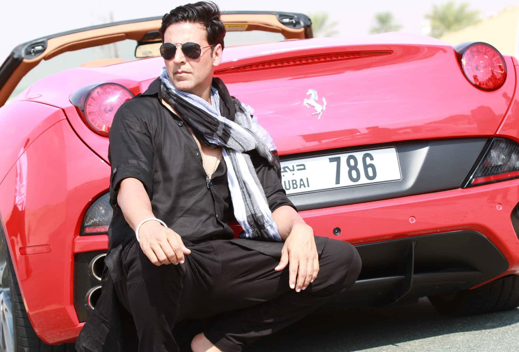 Akshay Kumar 桌布 壁紙 — 一名身穿黑色服裝和太陽眼鏡的男子自信地蹲在掛有杜拜車牌的鮮紅色法拉利開篷車旁。
