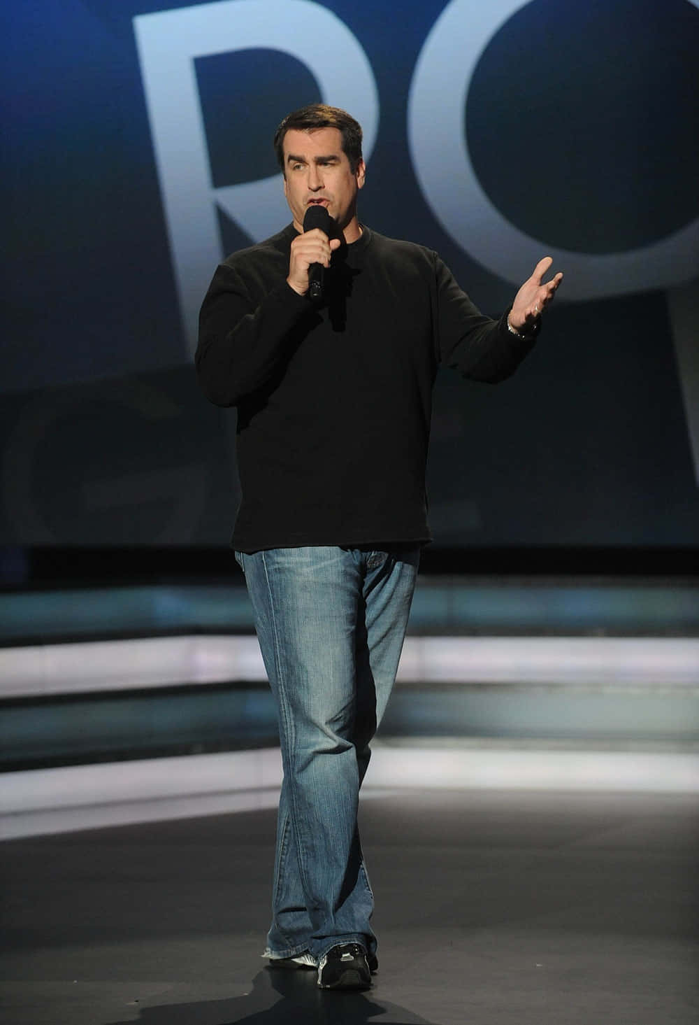 Rob Riggle 桌布 壁紙 — 一名身穿黑色毛衣和牛仔褲的男子在燈光昏暗的舞台上對著麥克風說話，身後有大型發光字母。