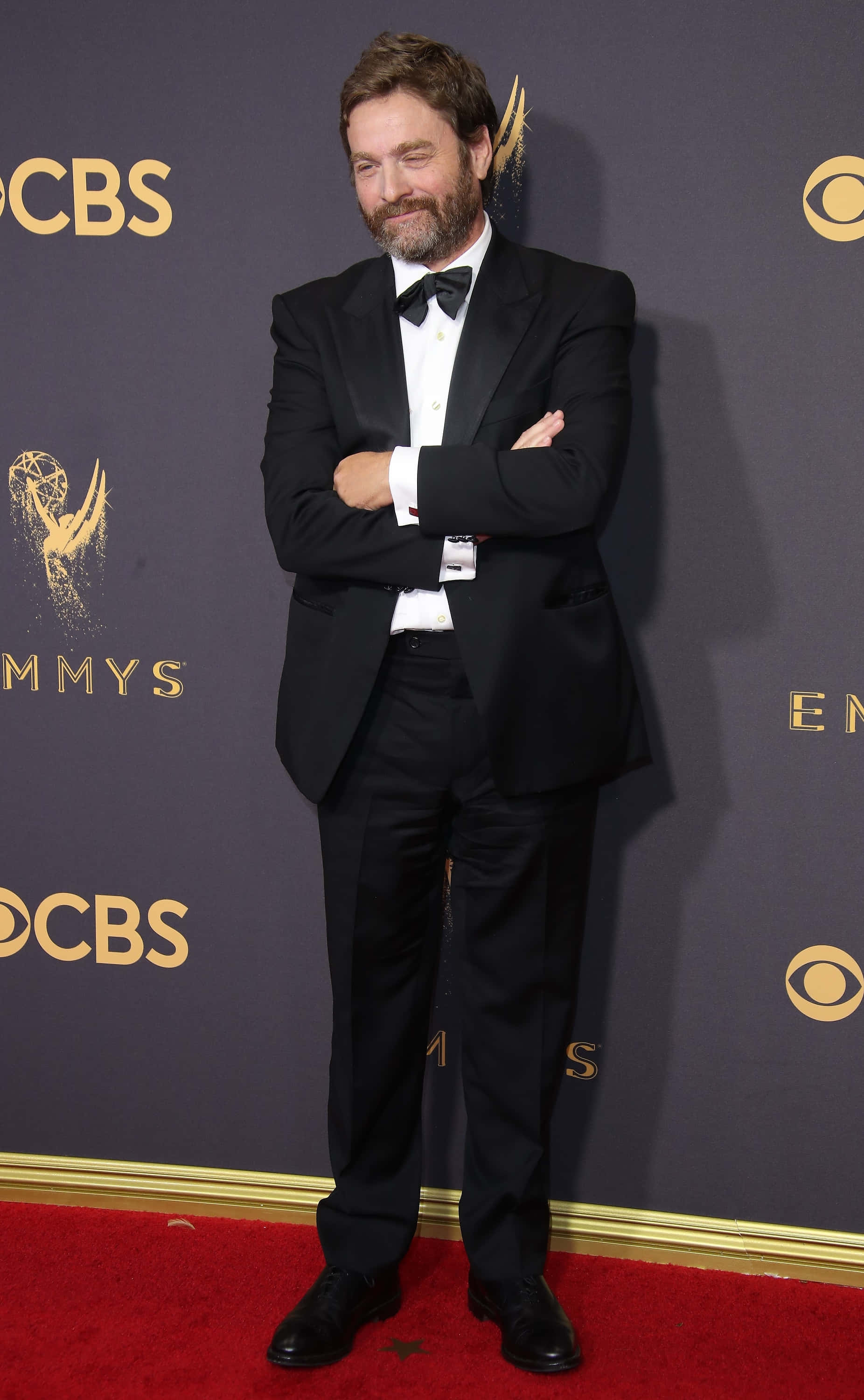 Zach Galifianakis 桌布 壁紙 — 一名身穿黑色燕尾服的男士在艾美獎活動的紅地毯上自信地交叉雙臂站立。