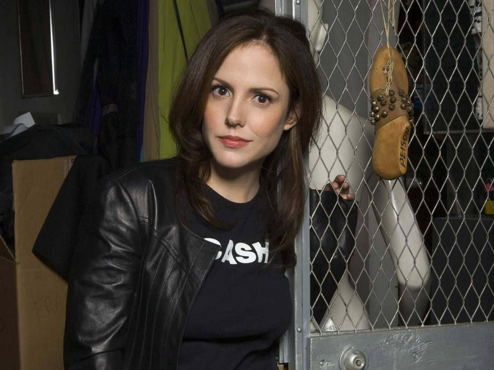 mary louise parker 桌布 壁紙 — 一名身穿黑色皮褸和T恤的女子站在雜亂的後台區域的鐵絲網圍欄後面。
