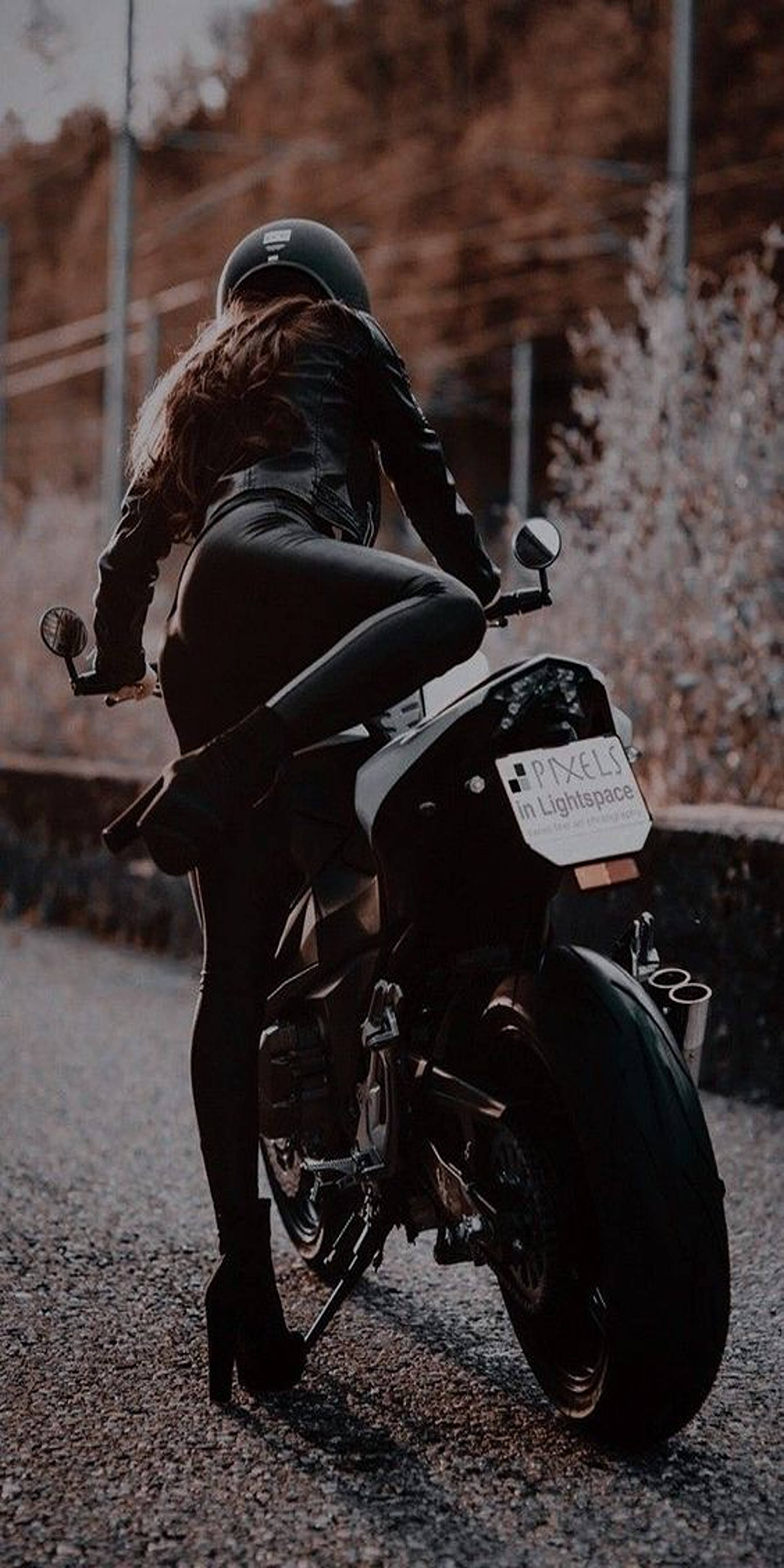 機車 桌布 壁紙 — 一名身穿黑色皮革和頭盔的女性，在碎石路上跨坐著一輛時尚電單車，背景是秋樹。