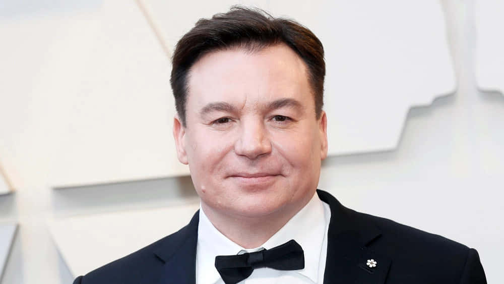 Mike Myers 桌布 壁紙 — 一名身穿黑色禮服和蝴蝶結的男子在白色幾何背景前對著鏡頭微笑。