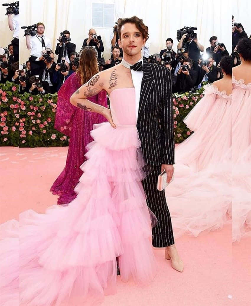 Met Gala 桌布 壁紙 — 一名身穿黑色細條紋西裝和粉紅色薄紗長裙的男子在粉紅色地毯上擺姿勢，周圍有攝影師和花卉佈置。