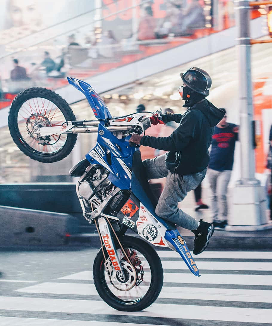 Dirtbike 桌布 壁紙 — 一名身穿黑色裝備的騎手，在模糊的行人和明亮燈光的背景下，騎著藍色越野車在城市的街道上大膽地表演翹前輪。