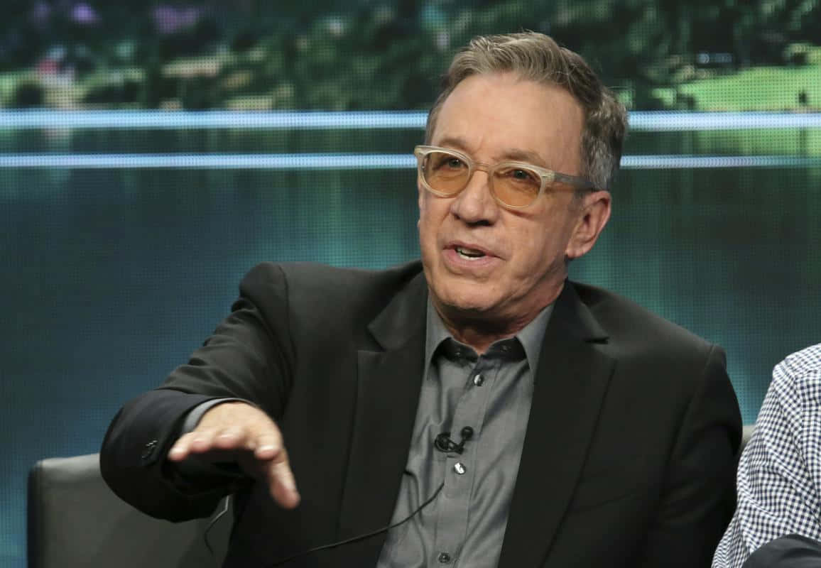 Tim Allen 桌布 壁紙 — 一名身穿黑色西裝、戴黃色眼鏡的男子在模糊綠色屏幕背景的座談會上發言時做手勢。