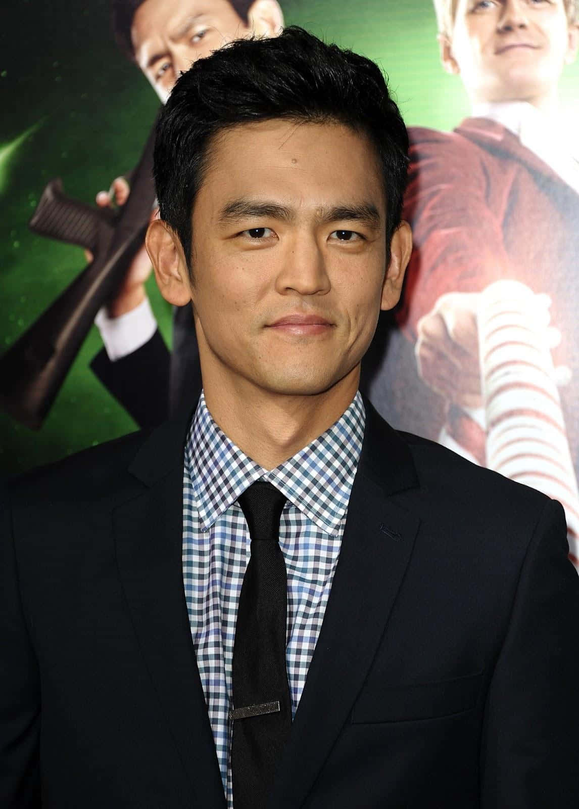 John Cho 桌布 壁紙 — 一名身穿黑色西裝和格仔恤衫的男子自信地站在電影首映禮上，身後有一張宣傳海報。