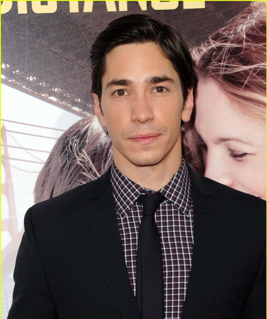 Justin Long 桌布 壁紙 — 一名身穿黑色西裝和格仔恤衫的男子站在以橋樑和女性面孔為背景的電影海報前。