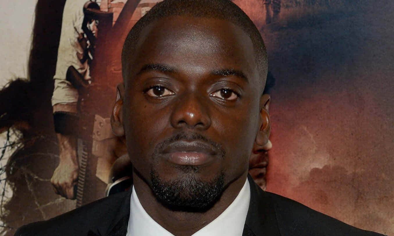 Daniel Kaluuya 桌布 壁紙 — 一名身穿黑色西裝和白色襯衫的男子，在電影海報背景前，表情嚴肅地直視鏡頭。