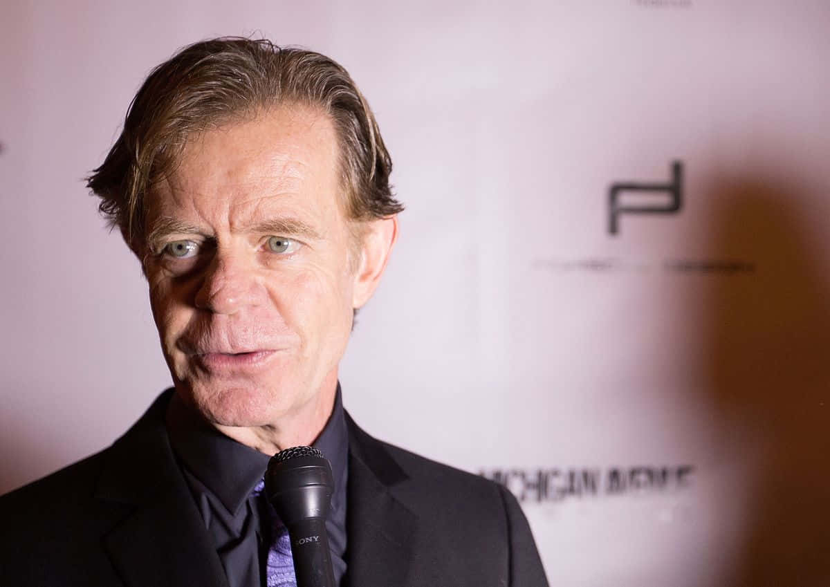 william h. macy 桌布 壁紙 — 一名身穿黑色西裝和紫色領帶的男子在紅地毯活動中對著麥克風說話，背景帶有品牌標誌。