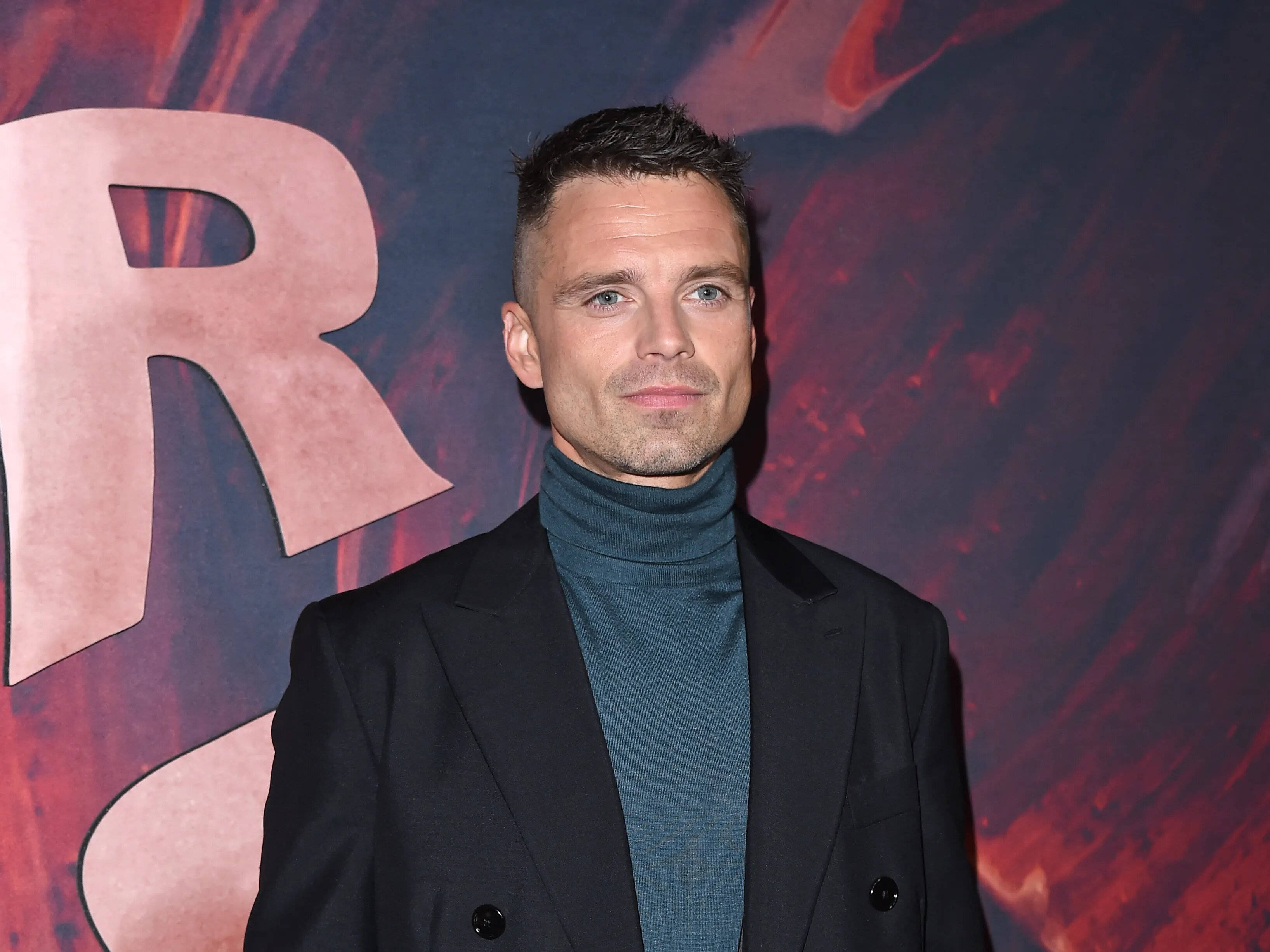 清新 桌布 壁紙 — 一名身穿黑色西裝外套和青綠色高領毛衣的男子自信地站在戲劇性的紅藍紋理背景前，背景上有一個大的粉紅色字母R。