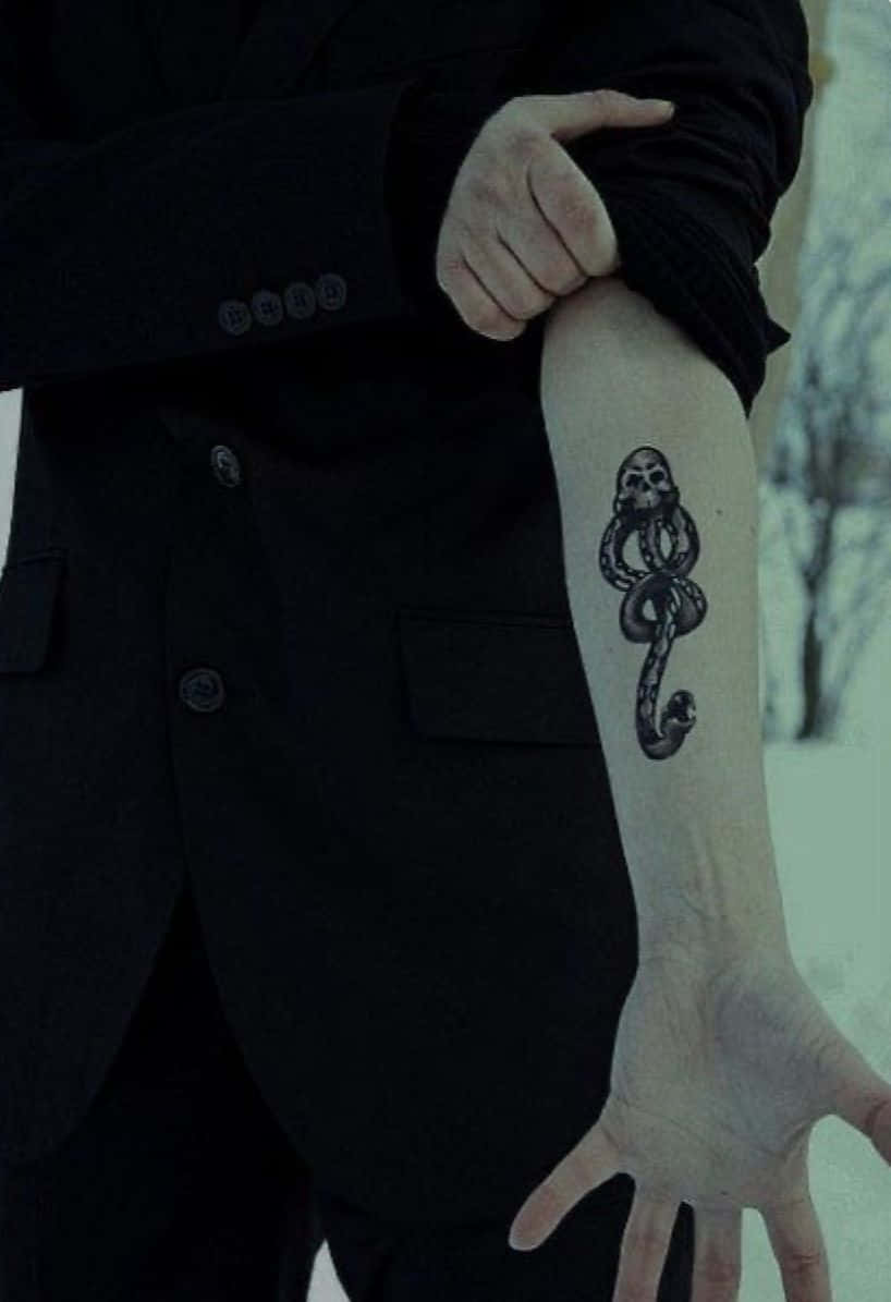Death Eaters 桌布 壁紙 — 一名身穿黑色西裝的人，在雪景背景下展示前臂上蛇纏繞骷髏的紋身。