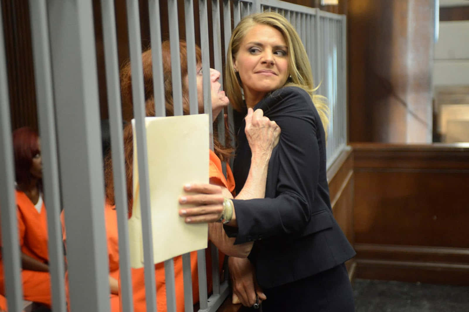 eliza coupe 桌布 壁紙 — 一名身穿黑色西裝的女子在法庭上透過金屬欄杆傾身，手持文件夾，而另一名身穿橙色囚服的女子站在她身後。