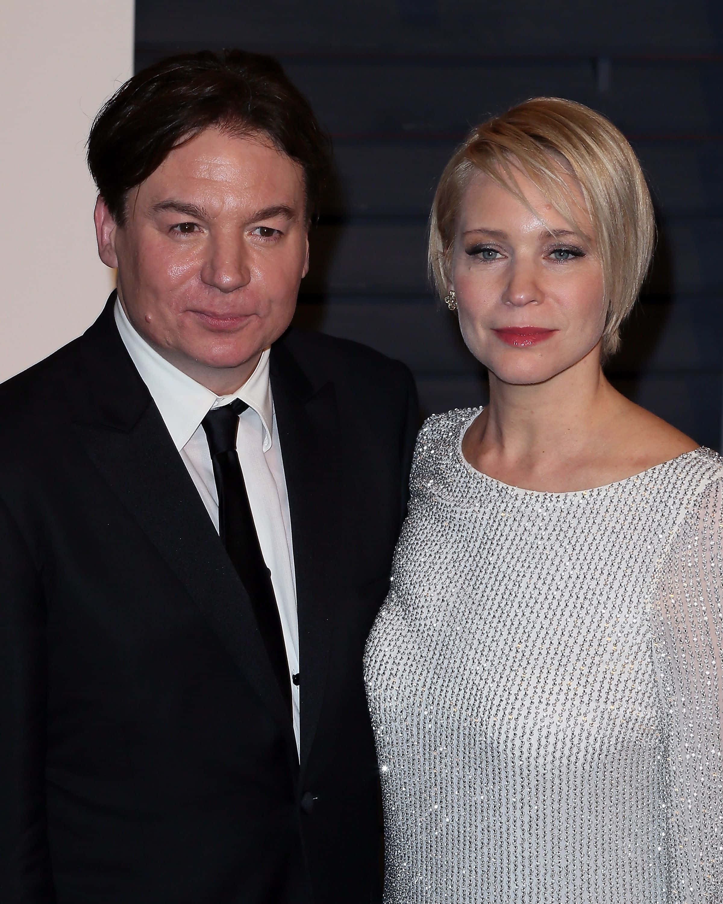 Mike Myers 桌布 壁紙 — 一名身穿黑色西裝的男子和一名身穿閃亮銀色連衣裙的女子在柔和燈光下於活動中一起擺姿勢。