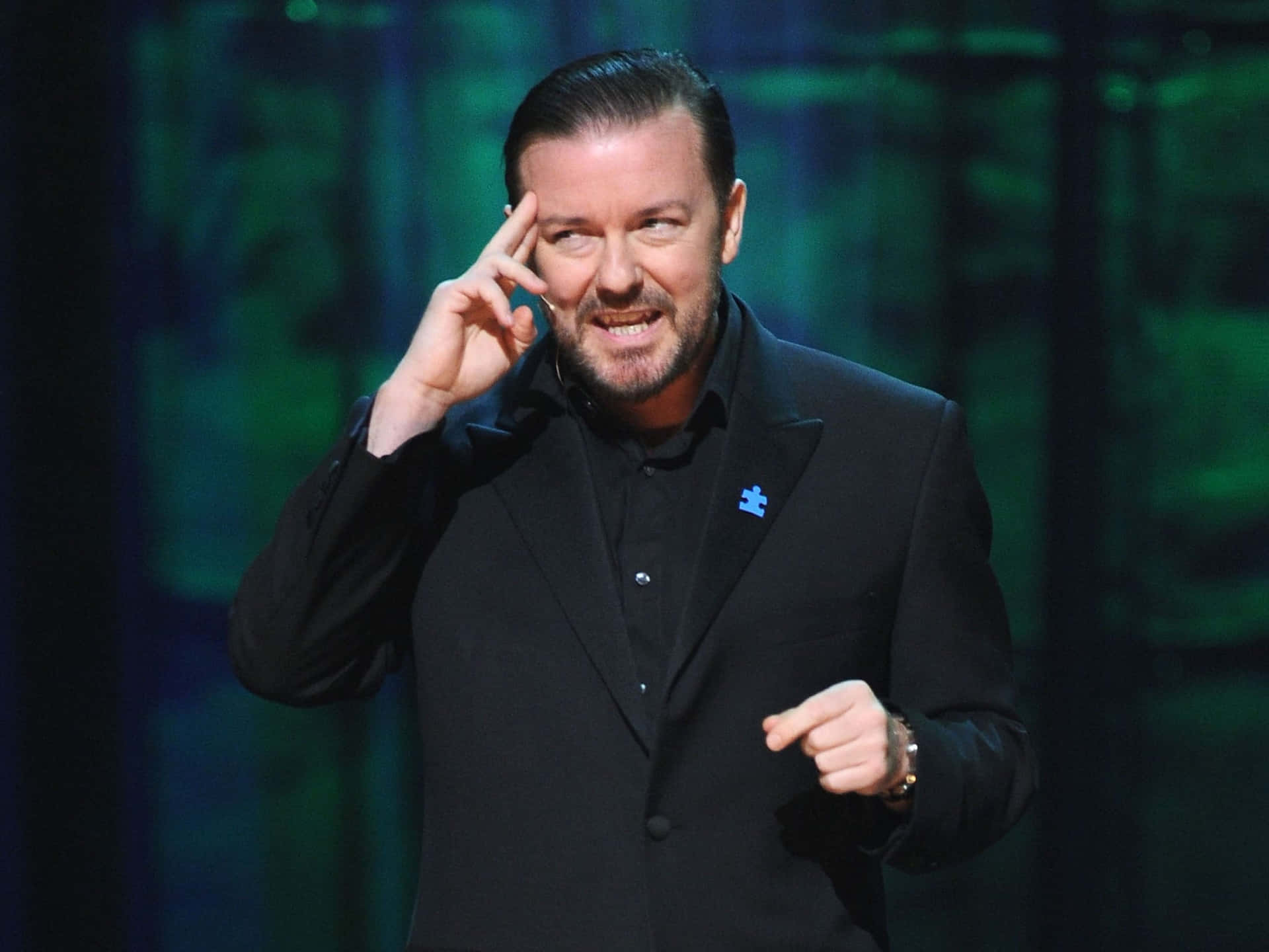 ricky gervais 桌布 壁紙 — 一名身穿黑色西裝的男子在昏暗的舞台上，在綠色背景燈光下生動地做手勢。