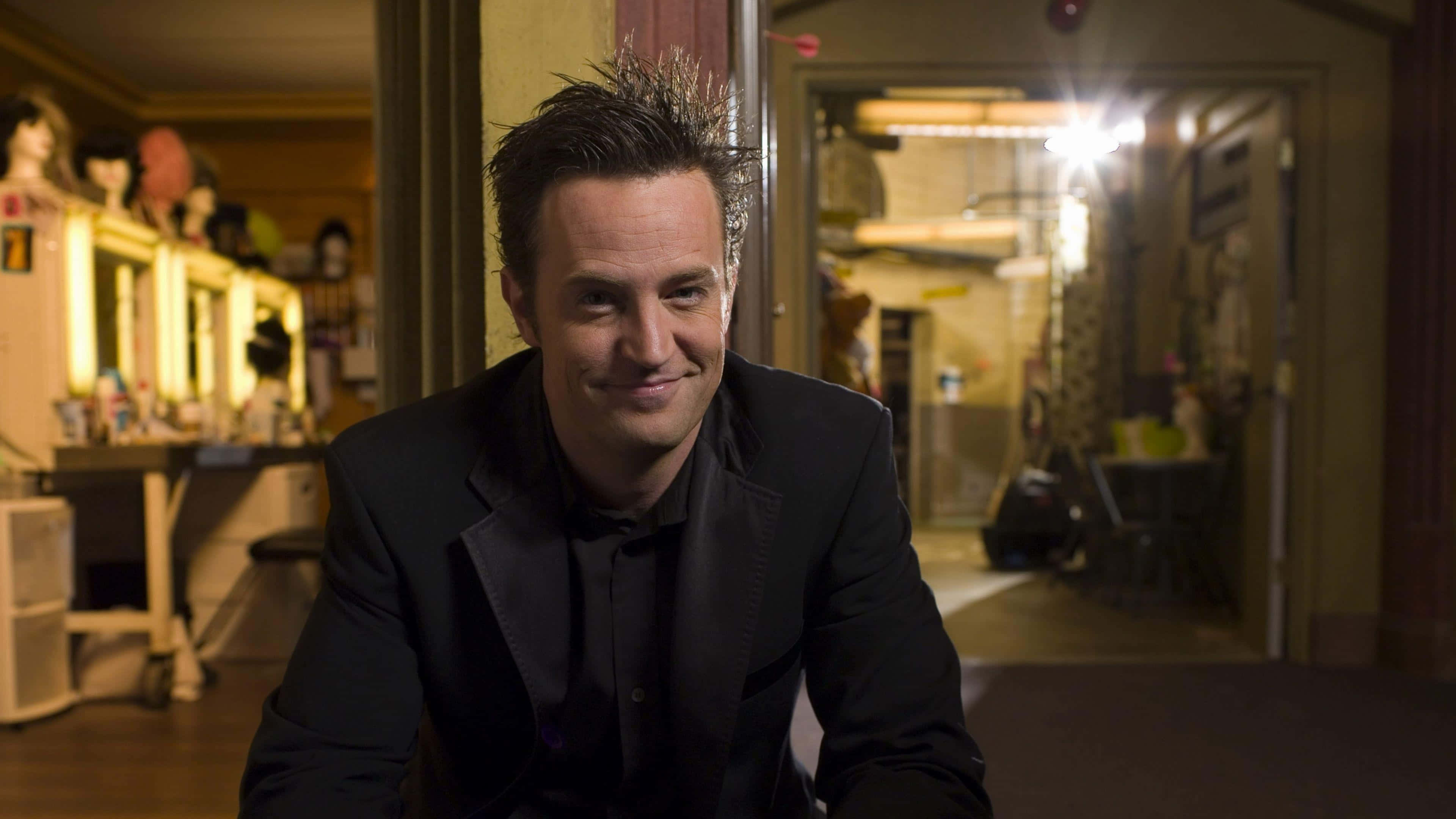 matthew perry 桌布 壁紙 — 一名身穿黑色西裝的男子在燈光昏暗的後台區域微笑，背景有化妝鏡和人體模型。