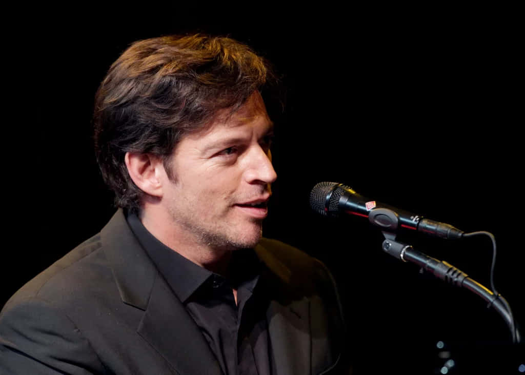 Harry Connick Jr 桌布 壁紙 — 一名身穿黑色西裝的男子在黑暗背景及聚焦舞台燈光下對著麥克風唱歌。