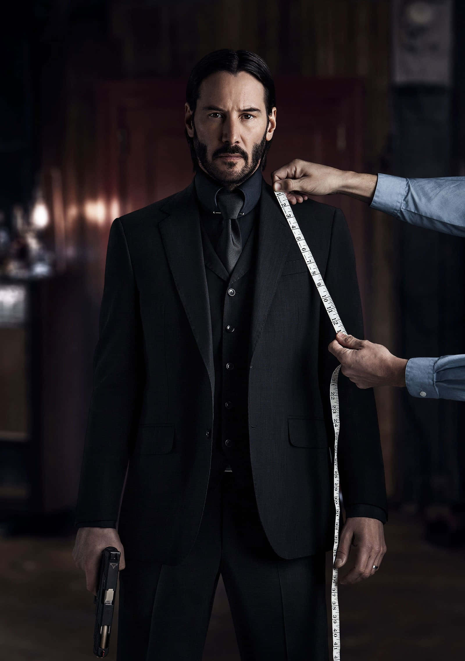 Keanu Reeves 桌布 壁紙 — 一名身穿黑色西裝的男子手持槍支，裁縫在昏暗的房間裡為他量度外套。