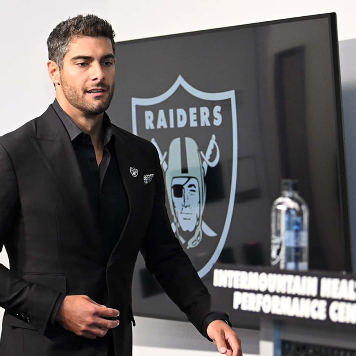 Jimmy Garoppolo 桌布 壁紙 — 一名身穿黑色西裝的男子自信地站在表演中心屏幕上嘅 Raiders 標誌前面。