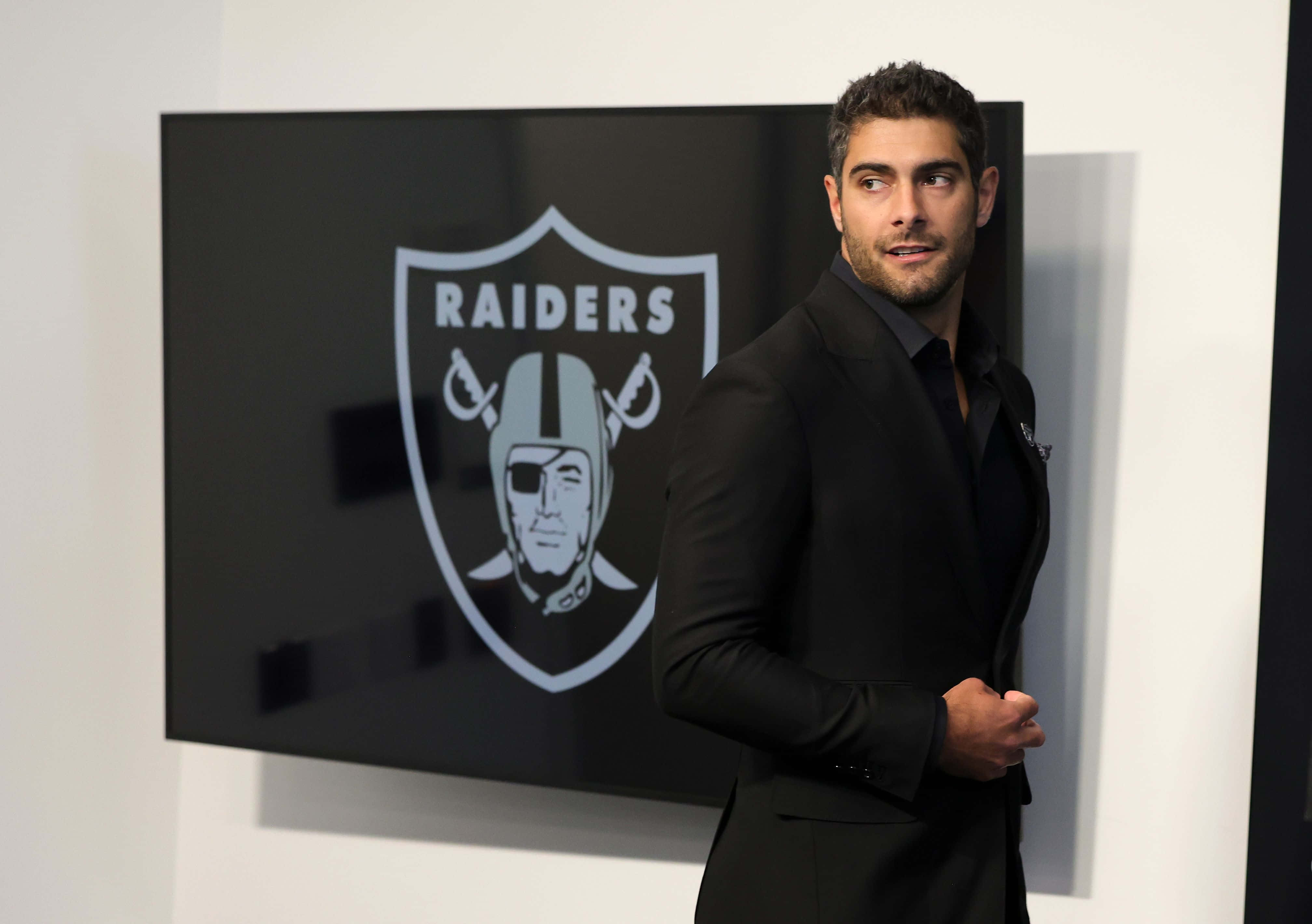 Jimmy Garoppolo 桌布 壁紙 — 一名身穿黑色西裝的男子自信地站在一個大型屏幕旁邊，屏幕上顯示著Raiders標誌，背景是簡約的室內環境。