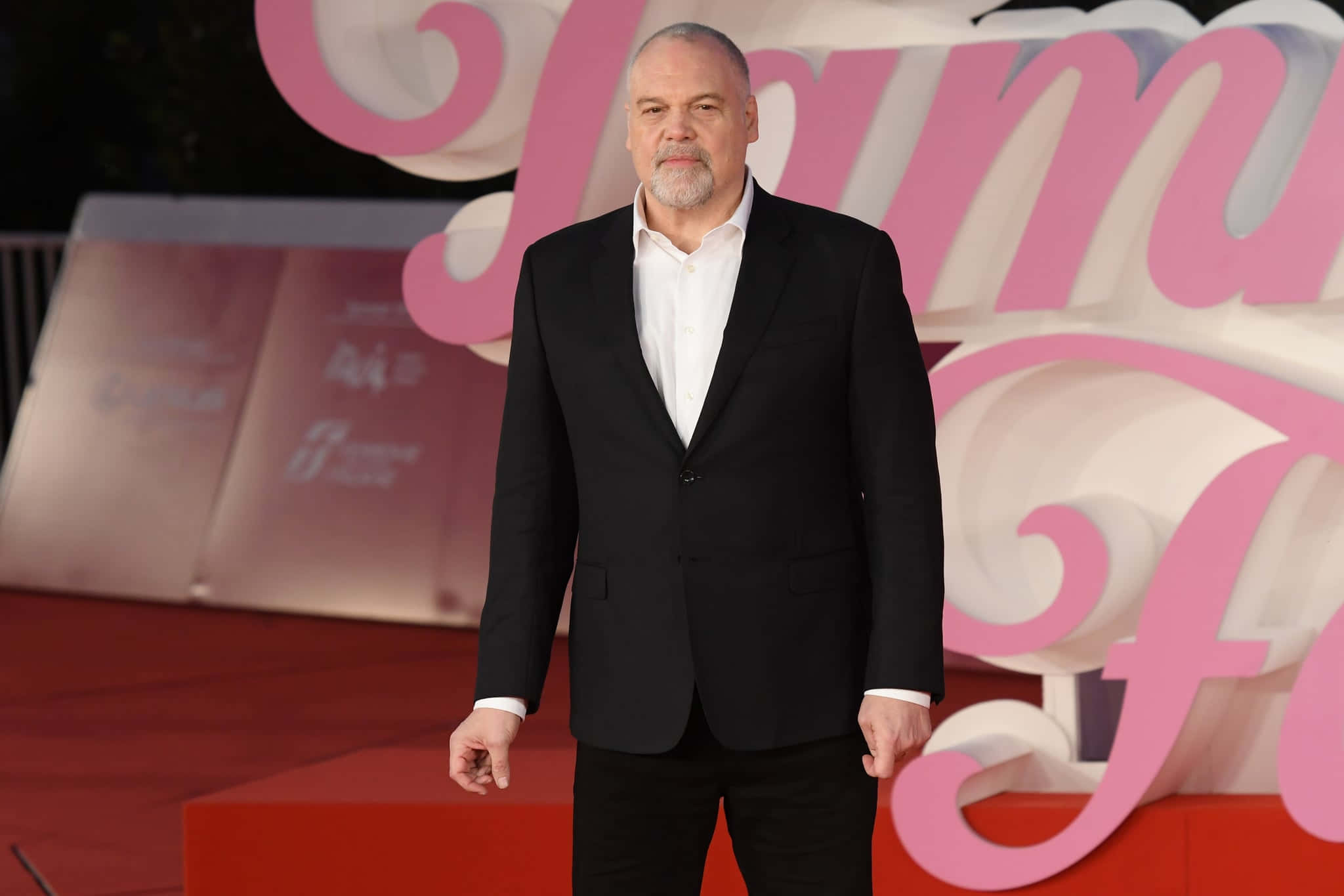 Vincent D'onofrio 桌布 壁紙 — 一名身穿黑色西裝的男子自信地站在紅地毯上，背景是大型粉白色活動佈景。