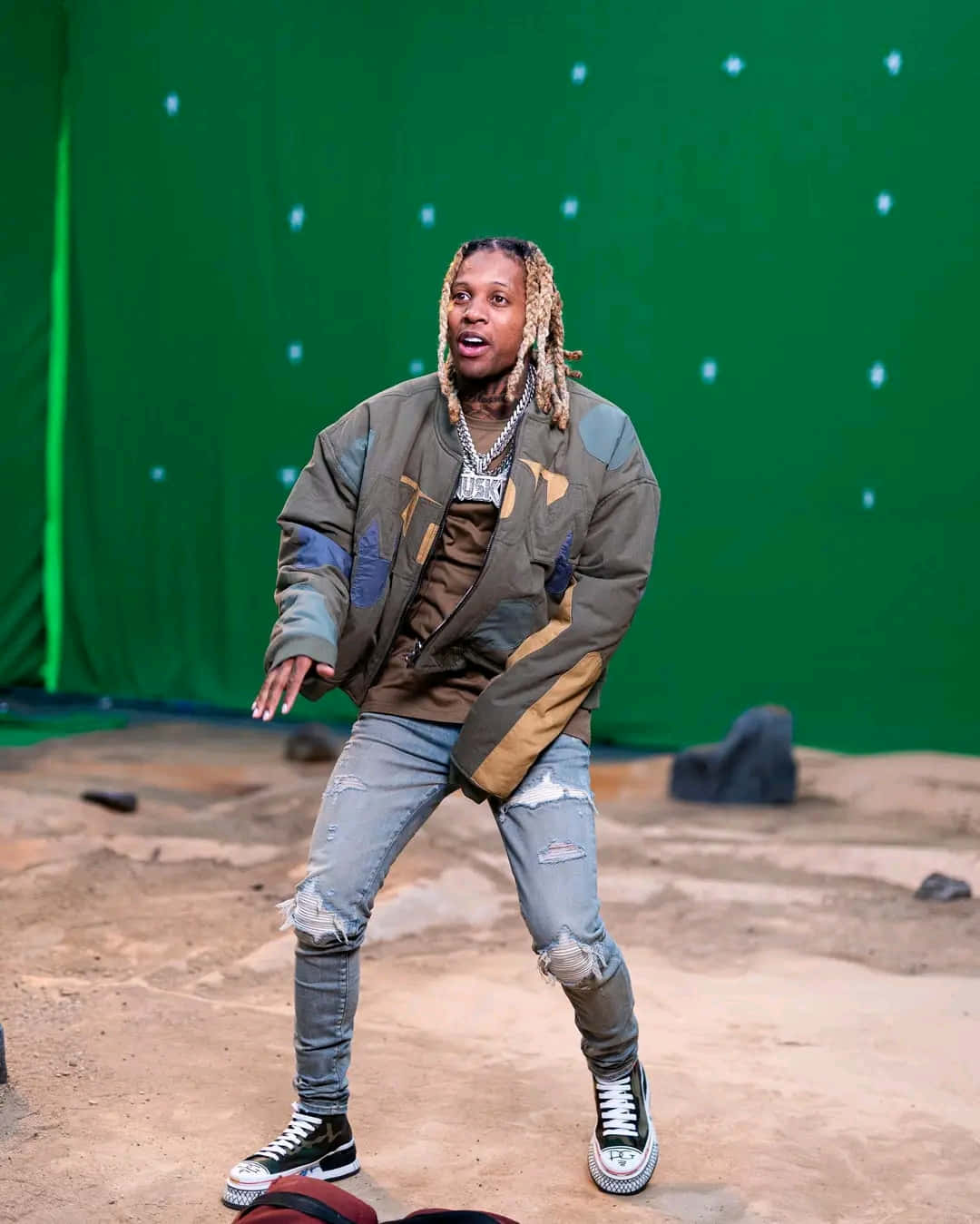 lil durk 桌布 壁紙 — 一名辮子頭髮的男子穿著補丁外套和破洞牛仔褲，在散落岩石和沙子的綠幕背景前動態擺姿勢。