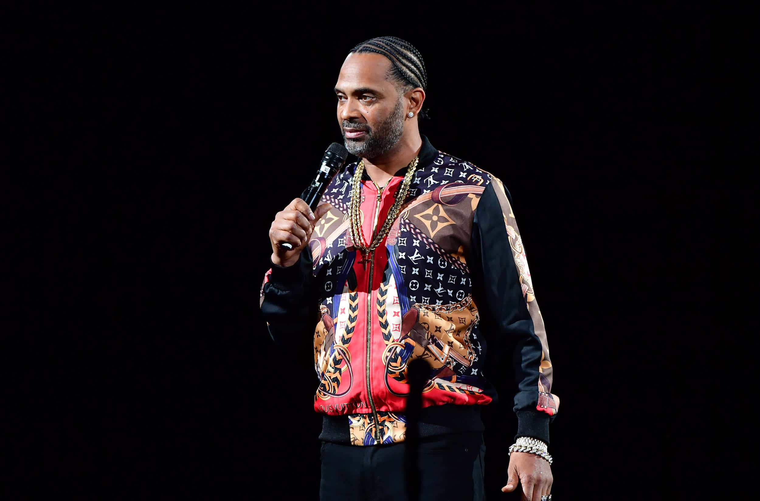 Mike Epps 桌布 壁紙 — 一名辮子髮型的男子站在黑暗的舞台上，手持麥克風，身穿色彩繽紛的圖案外套。