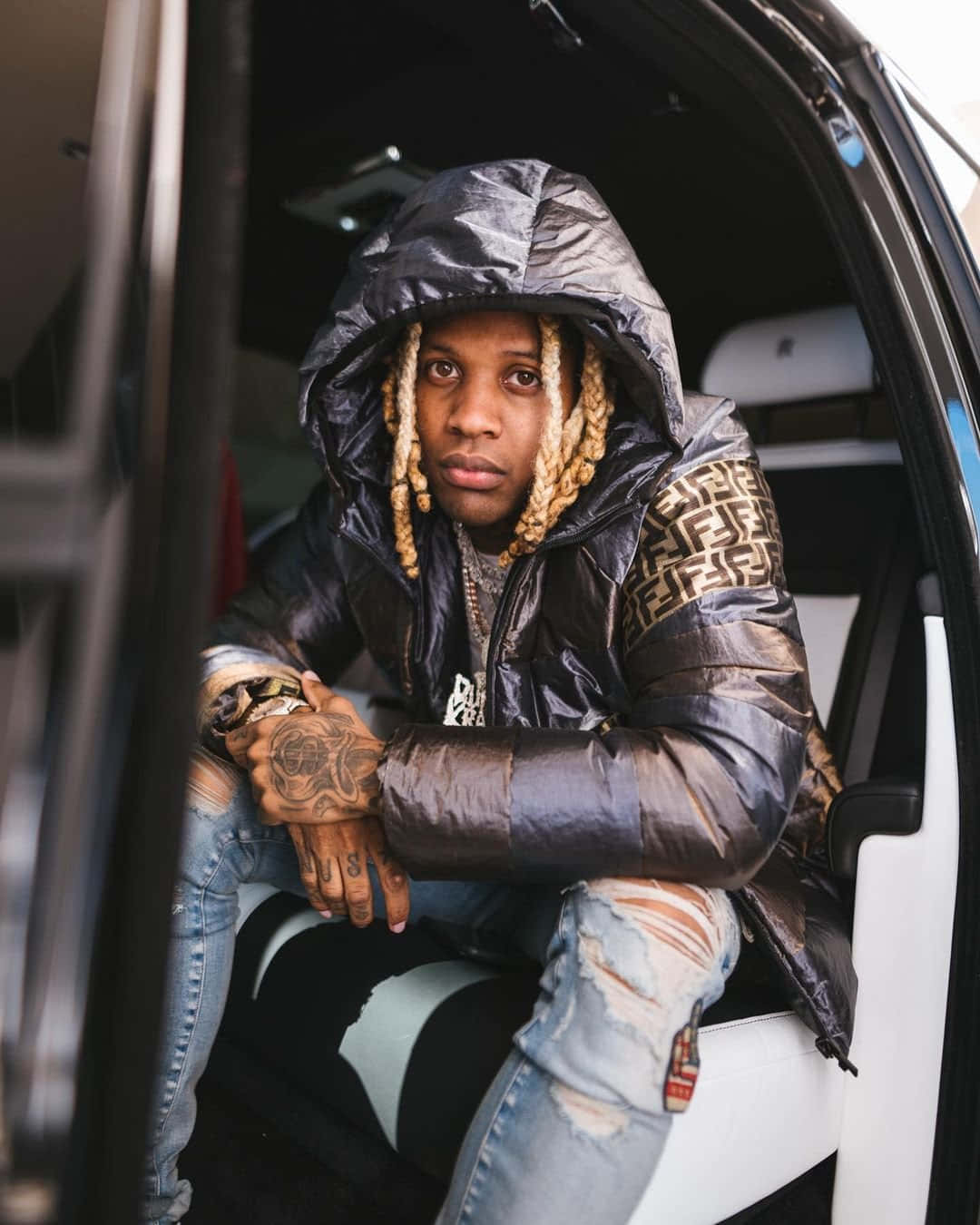 lil durk 桌布 壁紙 — 一名辮髮且有紋身的男子坐在車內，身穿帶有金色標誌袖子的閃亮黑色羽絨外套。