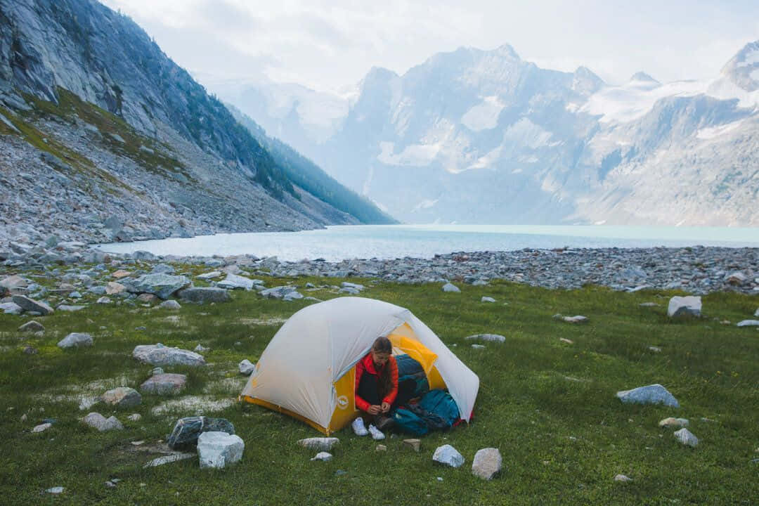Winter Camping 桌布 壁紙 — 一名遠足者坐在湖邊草地上一個黃白相間的帳篷內，背後是白雪覆蓋的山峰。