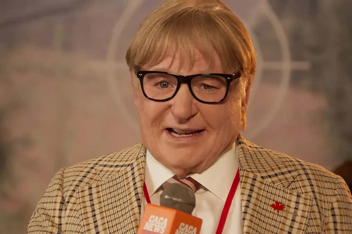 Mike Myers 桌布 壁紙 — 一名金髮戴黑框眼鏡的男子，在模糊背景前微笑，手持印有「CACA NEWS」的咪高峰。