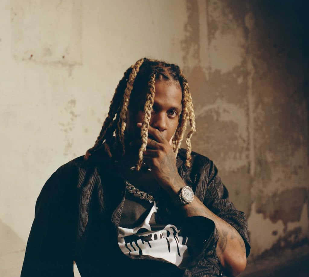 lil durk 桌布 壁紙 — 一名金髮辮子、有紋身的男子，在風化的牆壁前沉思擺姿勢，手靠近嘴邊，身穿黑色外套和骷髏圖案T恤。