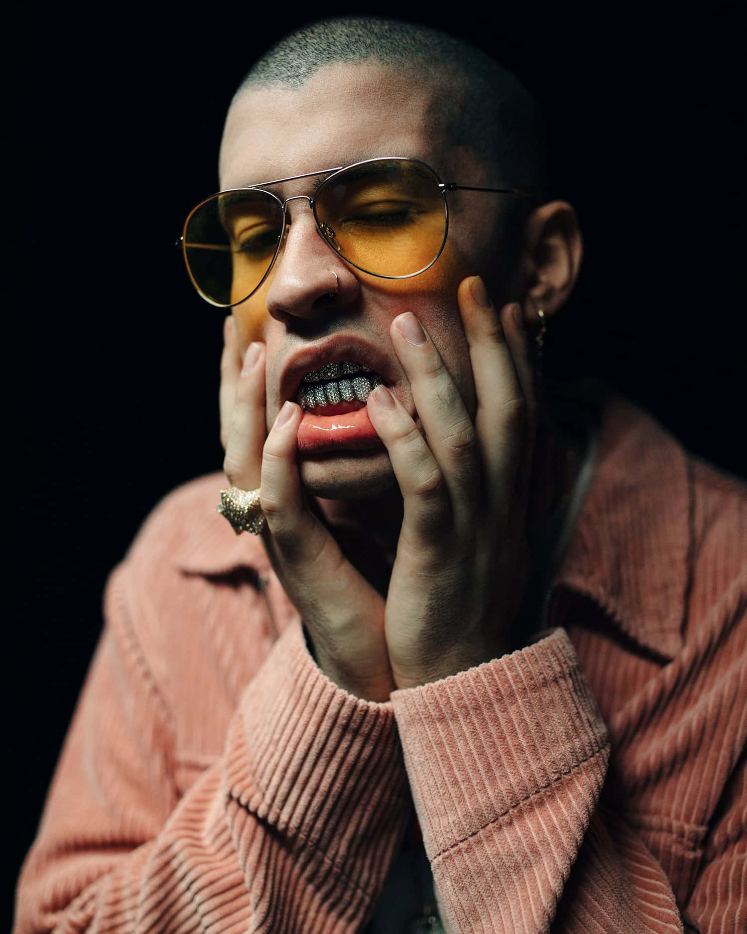 Bad Bunny 桌布 壁紙 — 一名鑲有鑽石牙齒、戴著黃色飛行員眼鏡的男子，在黑色背景前將雙手按在臉上。
