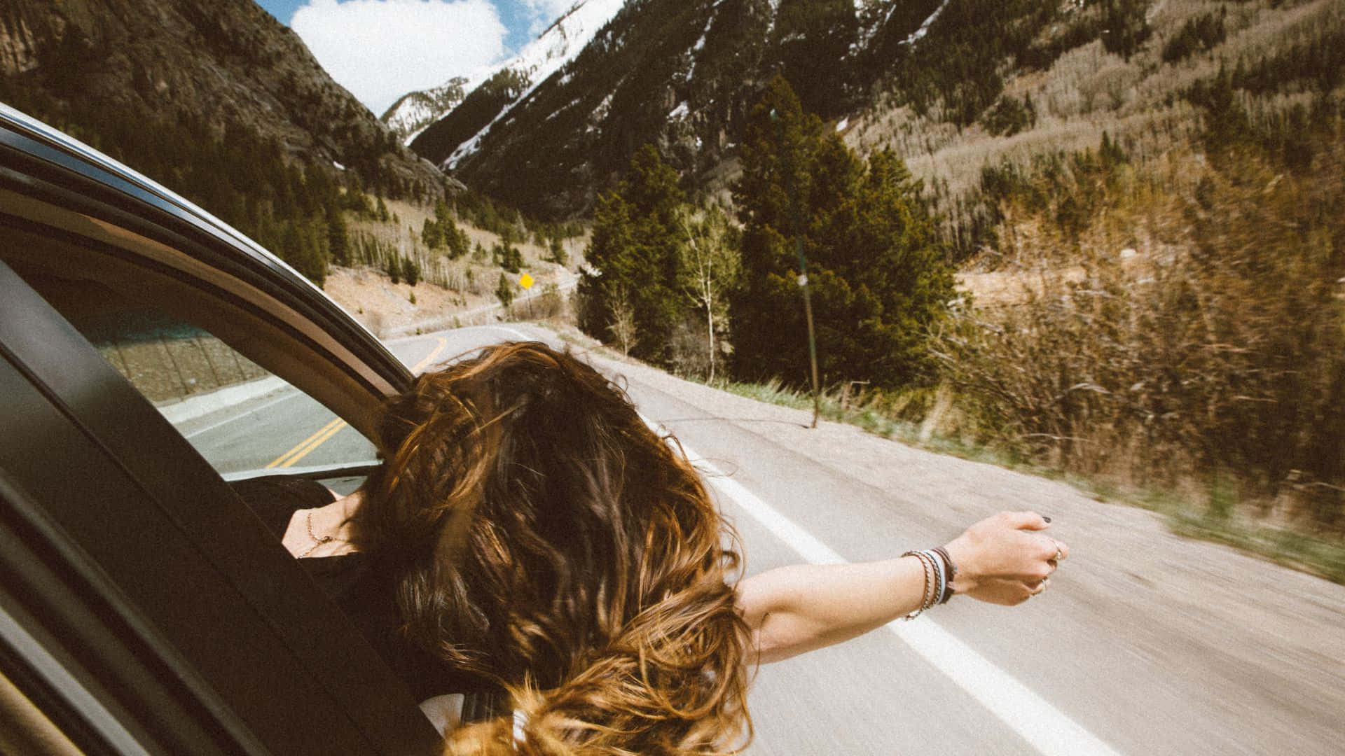 Beautiful Travel 桌布 壁紙 — 一名長髮人士從行駛中的汽車車窗探出身子，雙臂張開，車輛沿著蜿蜒的山路行駛，兩旁是常綠森林和白雪覆蓋的山峰，天空晴朗。