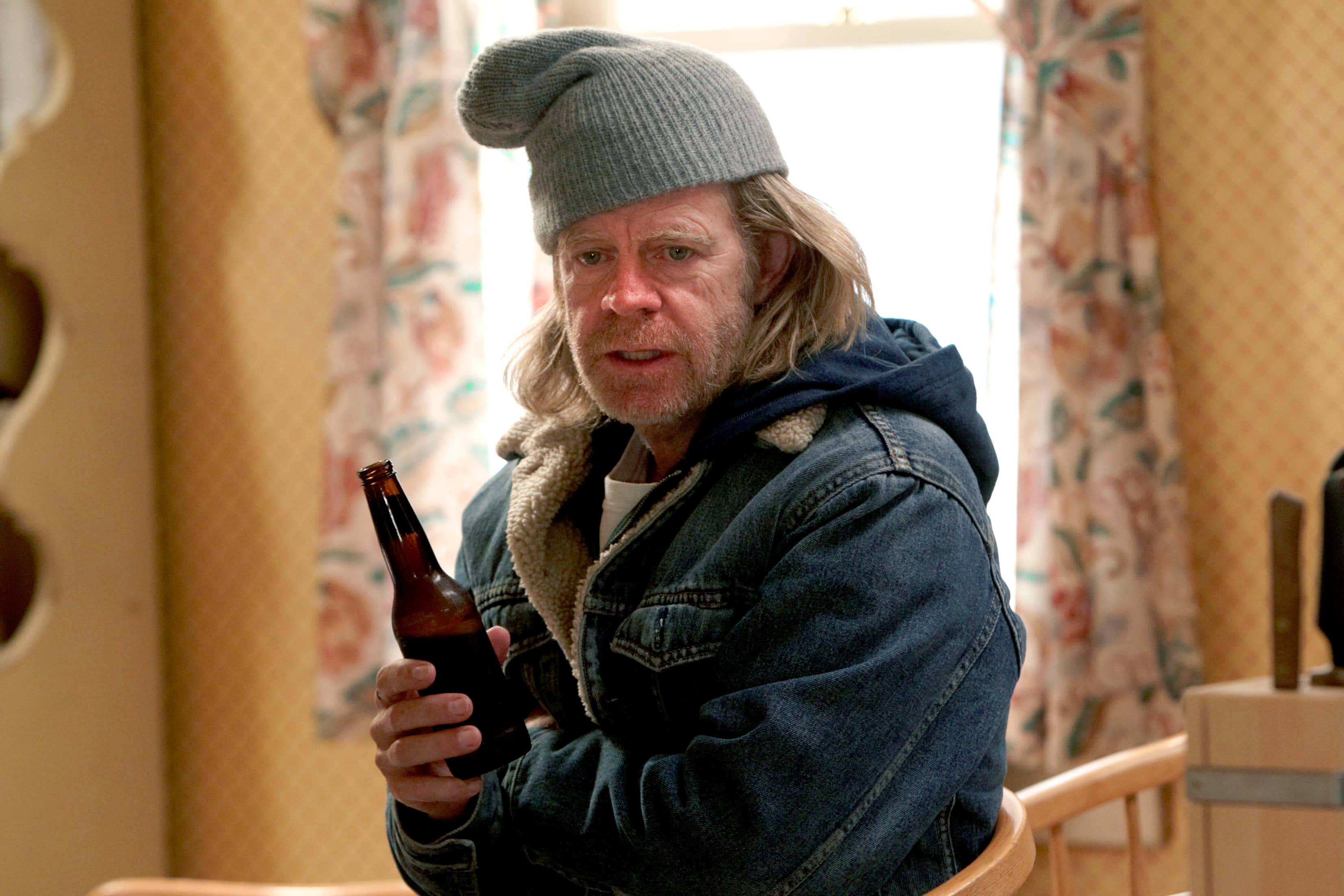 william h. macy 桌布 壁紙 — 一名長髮戴灰色冷帽的男子，坐在室內花卉窗簾旁，手持一個空啤酒瓶。