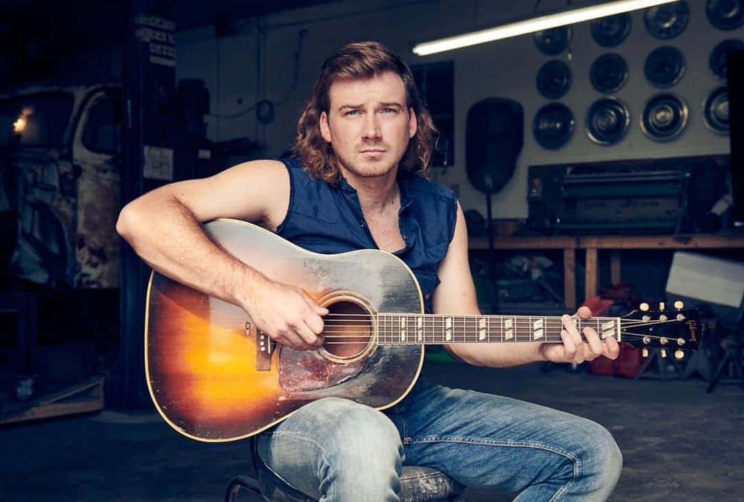Morgan Wallen 桌布 壁紙 — 一名長髮男子坐在昏暗的車庫中，在螢光燈下拿著一把旭日紋原聲結他。