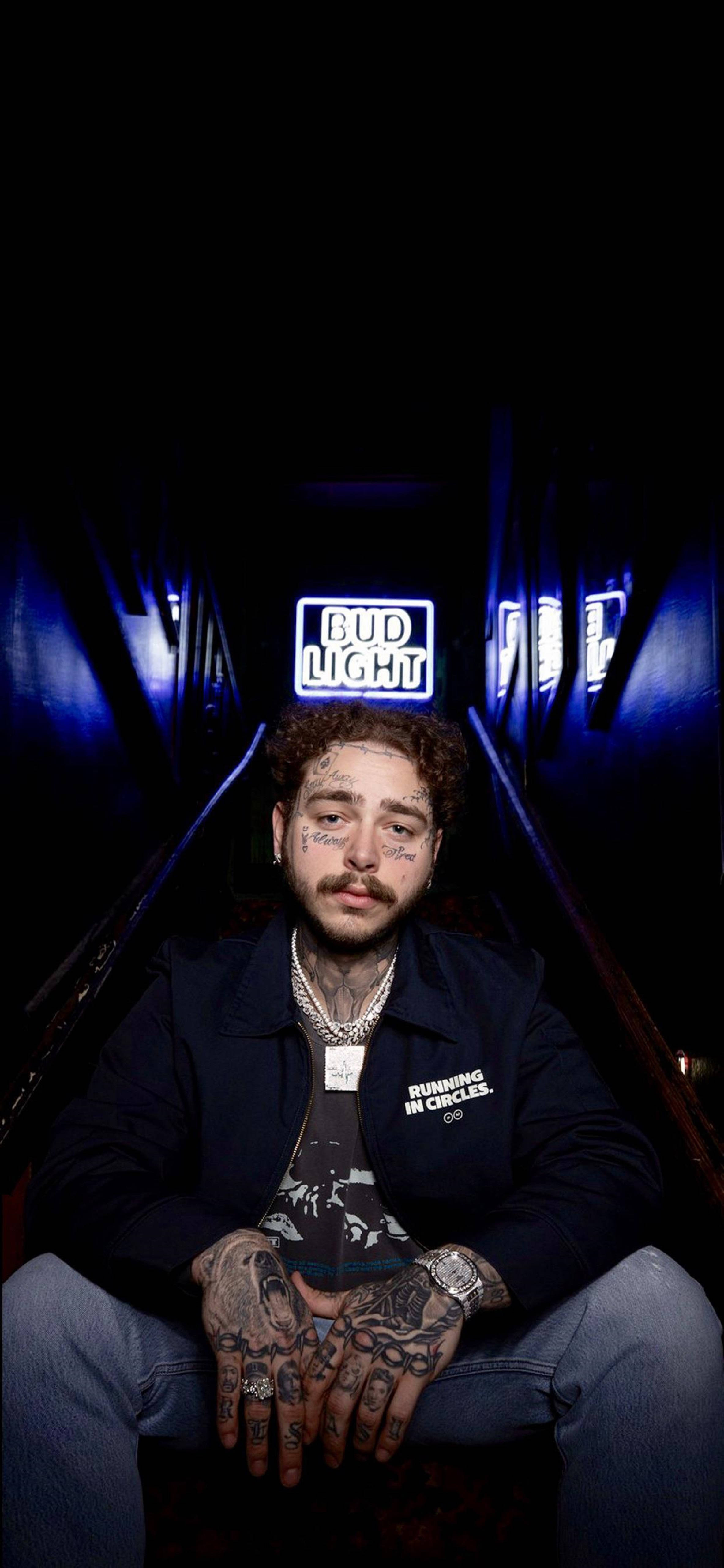 Post Malone Bud Light Merch 壁紙 — 一名面部有紋身的男子坐在黑暗走廊中央，被發出藍光的Bud Light標誌照亮。