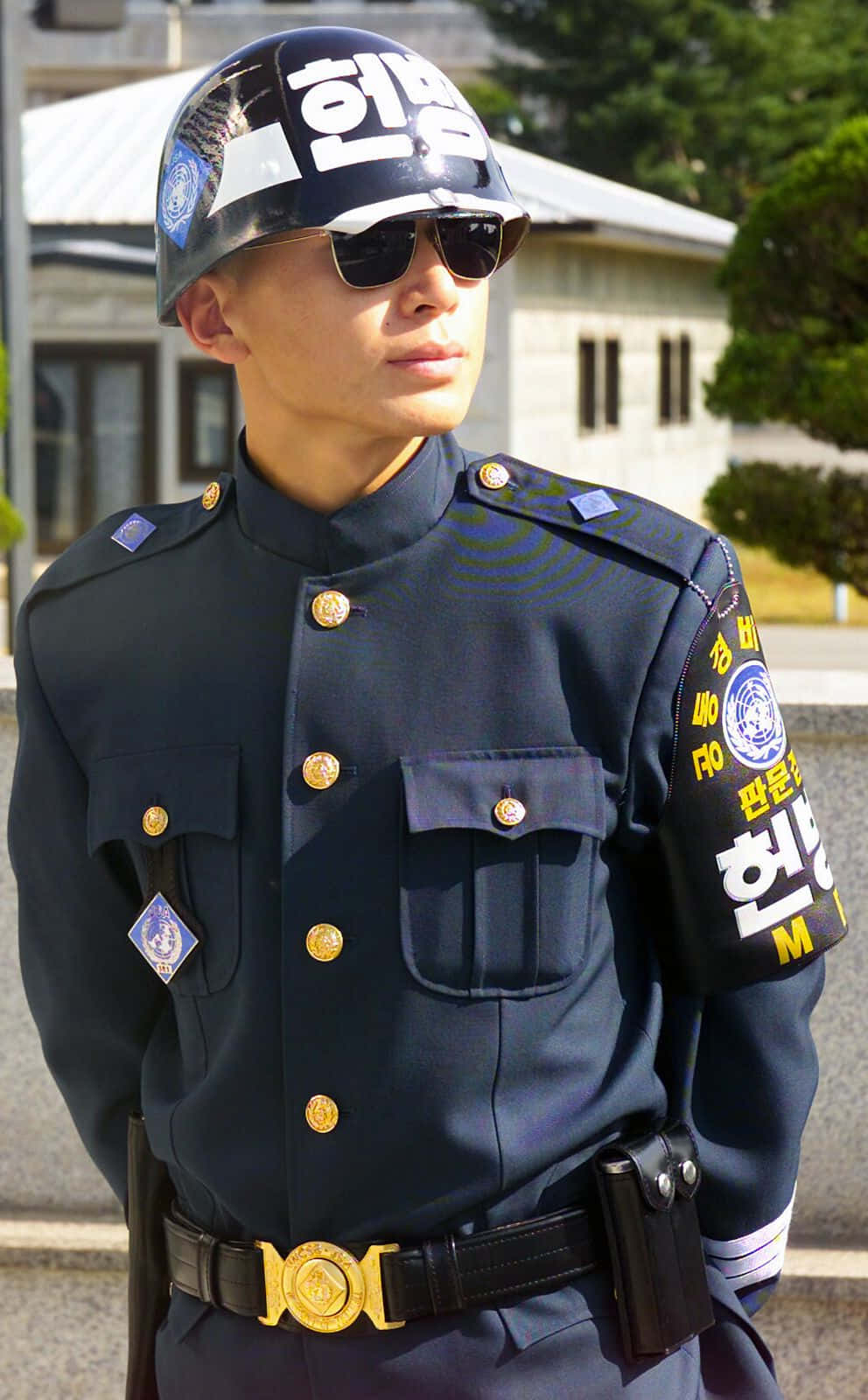 警察 桌布 壁紙 — 一名韓國警察穿著深色制服和頭盔，在明亮的陽光下警覺地站在戶外。