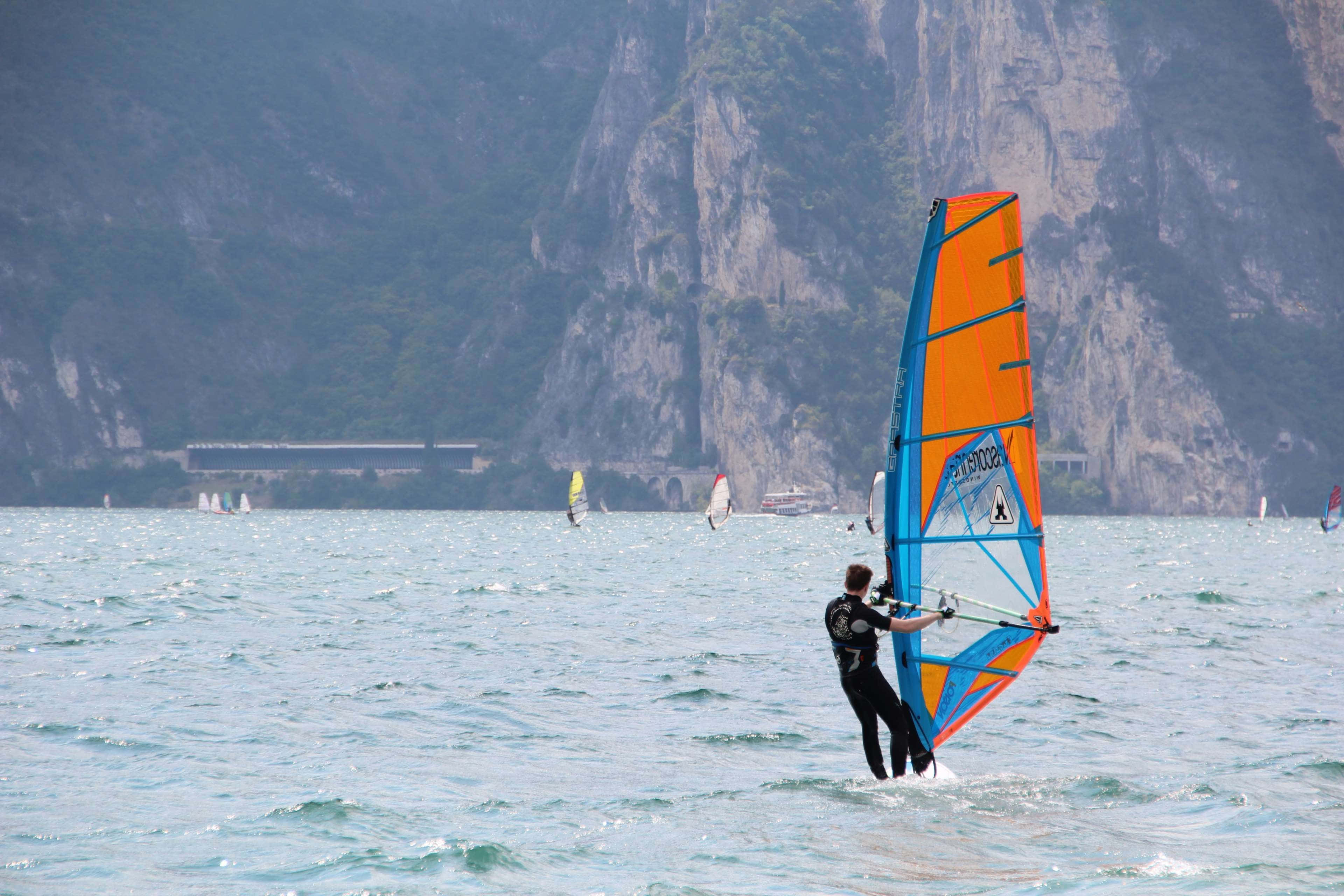 Beach Water Sports 桌布 壁紙 — 一名風帆選手在陽光明媚的湖面上滑行，配以鮮豔的橙色和藍色風帆，背景是高聳的綠色山脈。