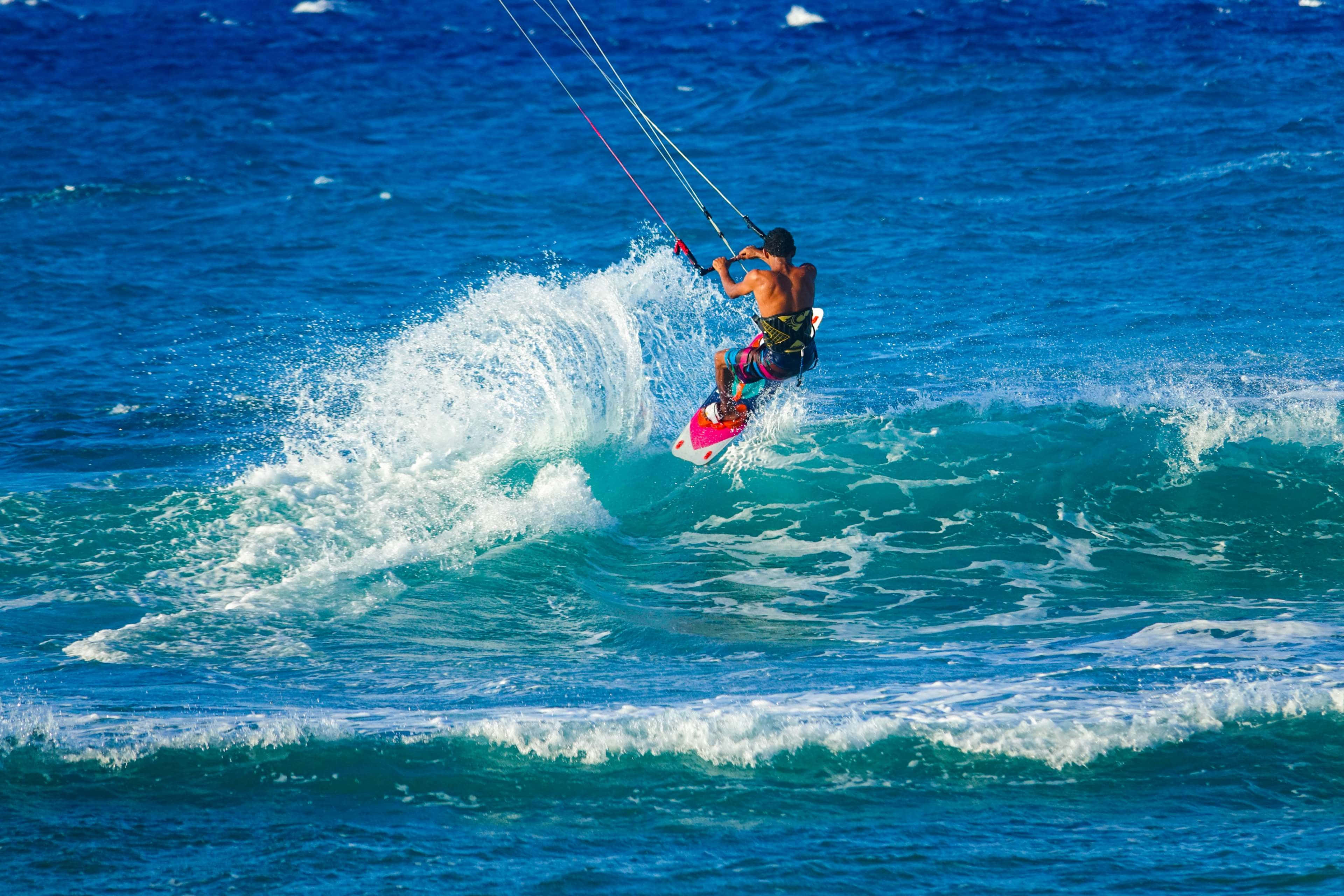 Beach Water Sports 桌布 壁紙 — 一名風箏衝浪者在明亮的藍天下，騎乘著藍綠色的海浪，激起動態的水花。
