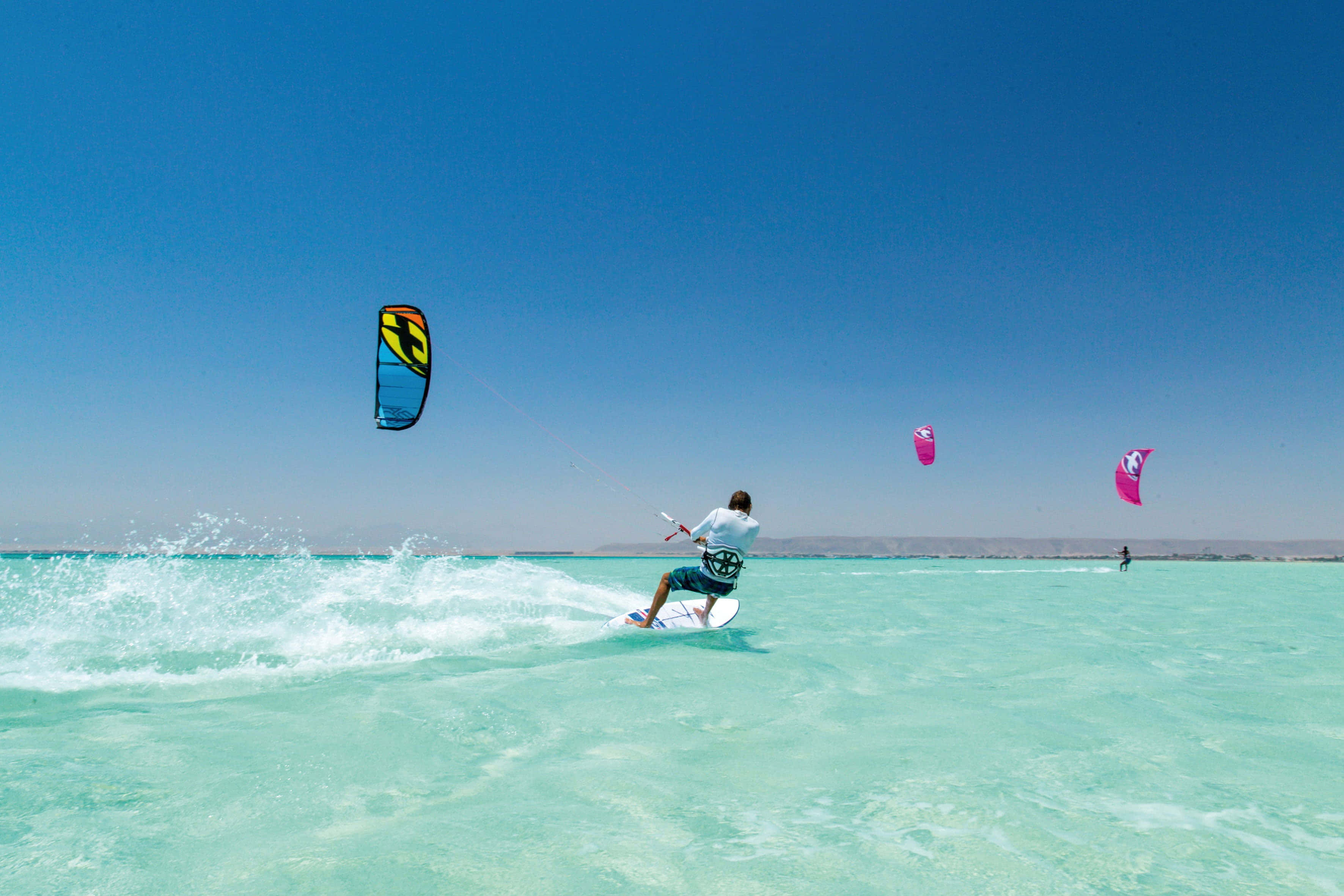 Beach Water Sports 桌布 壁紙 — 一名風箏衝浪者在晴朗藍天下乘著碧綠海浪，色彩繽紛的風箏在頭頂翱翔。