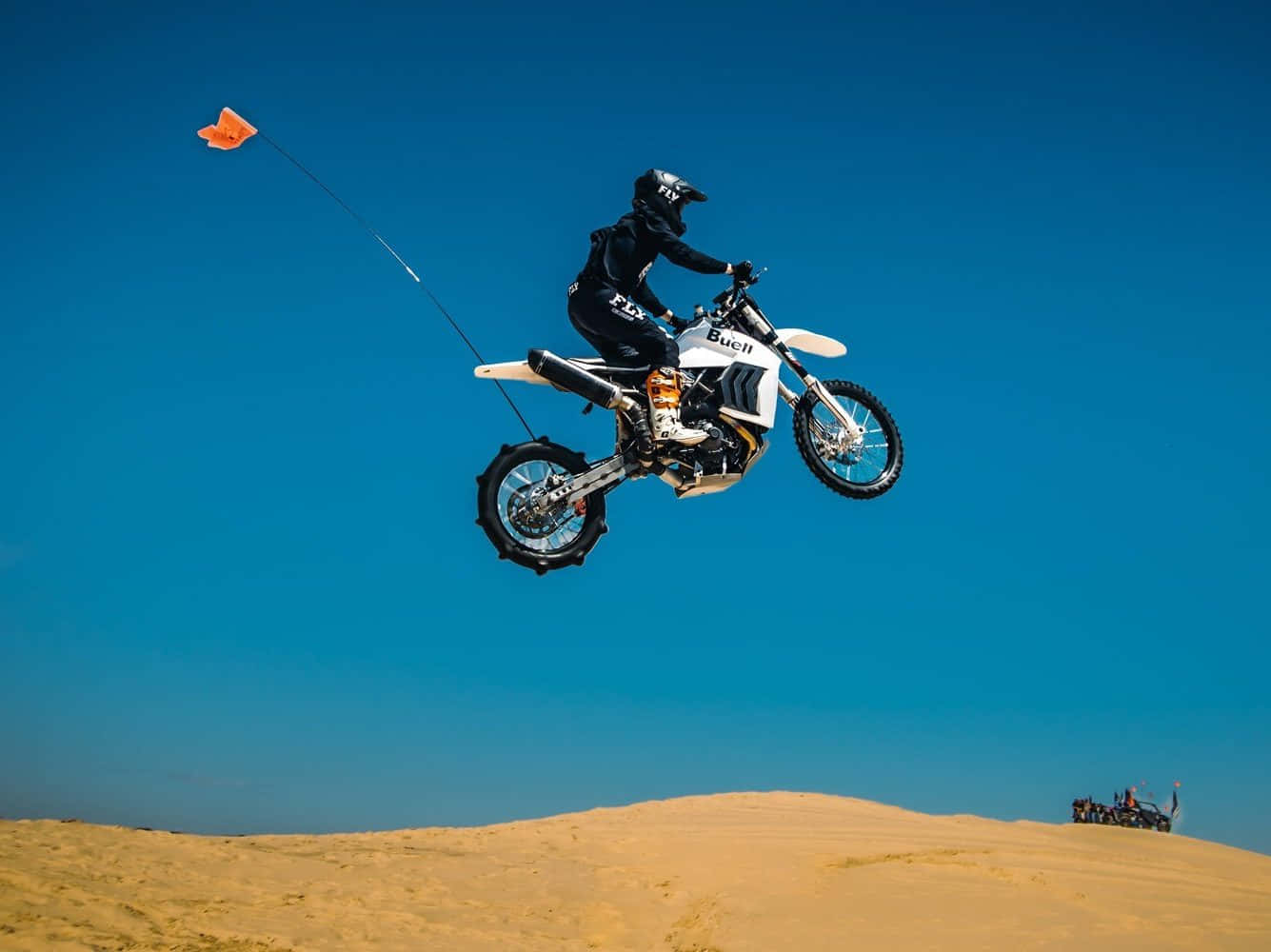 Dirtbike 桌布 壁紙 — 一名騎手騎著白色越野摩托車在金色沙丘上空飛翔，藍天白雲下，橙色旗幟在身後飄揚。