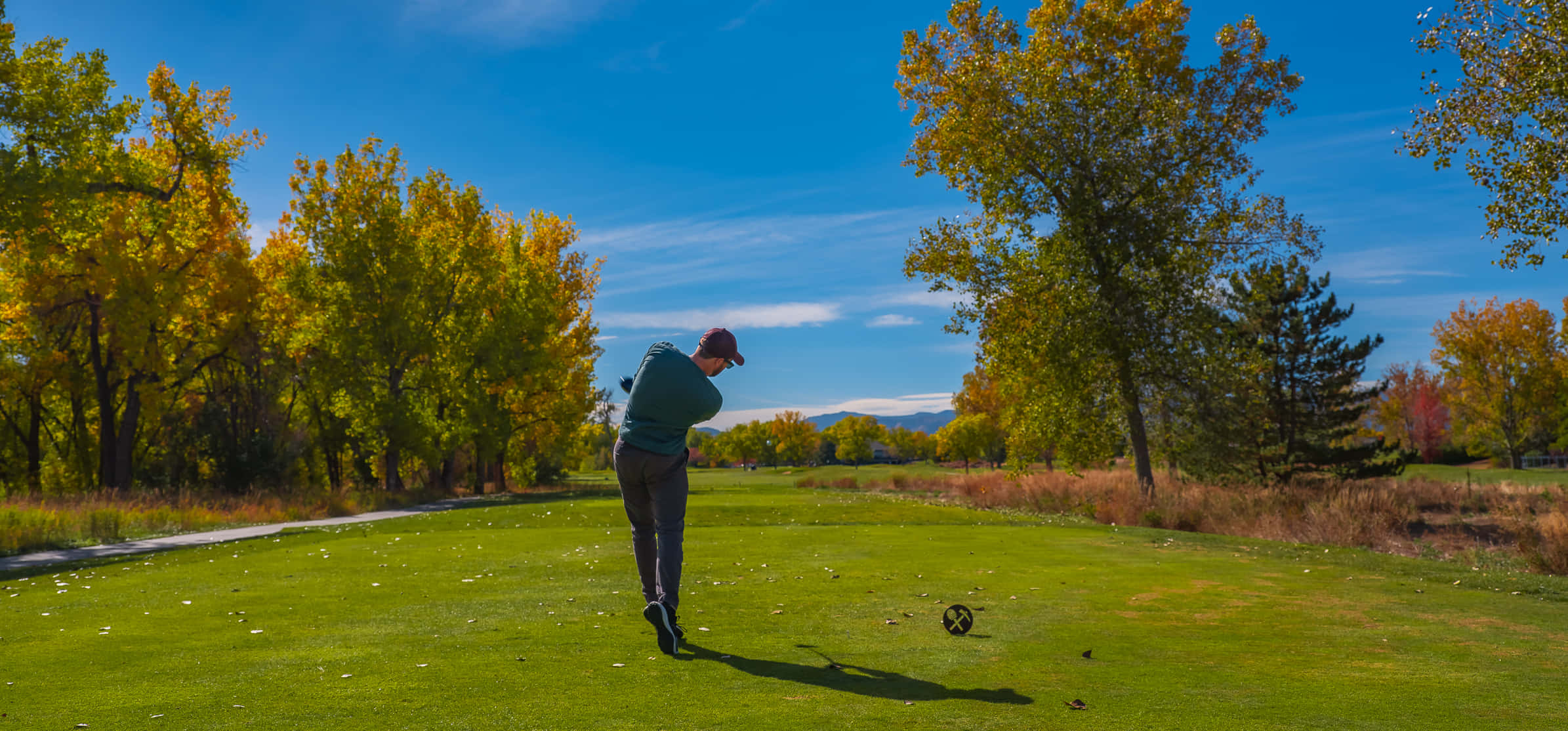 Golf Green 桌布 壁紙 — 一名高爾夫球手在晴朗藍天下，於金黃秋樹環繞的翠綠球道上揮桿擊球。